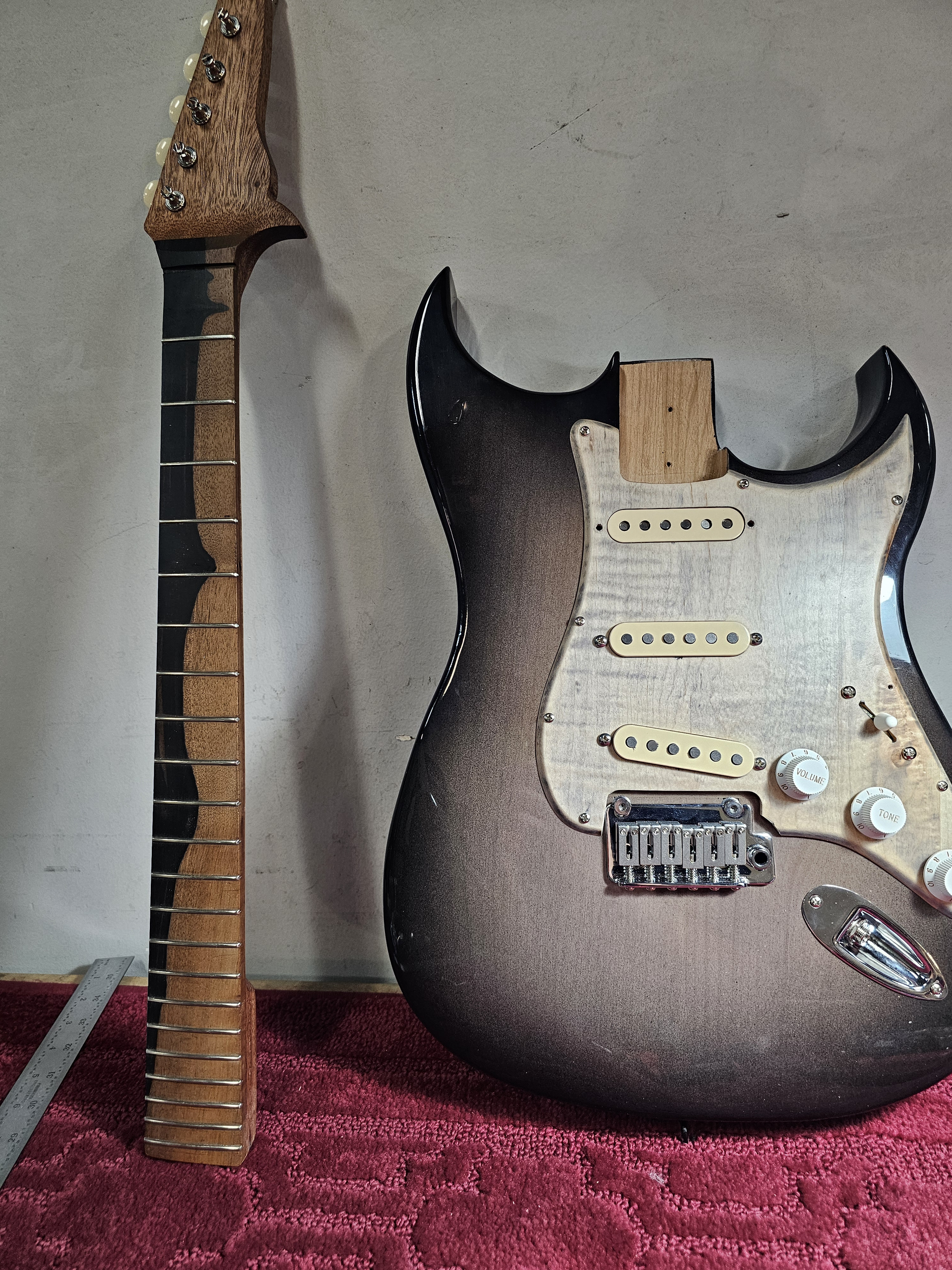 Disassembled electric guitar with a wooden neck, tuning pegs, and fretboard on the left; the guitar body, pickups, volume and tone knobs, and bridge on the right, placed on a red textured surface.