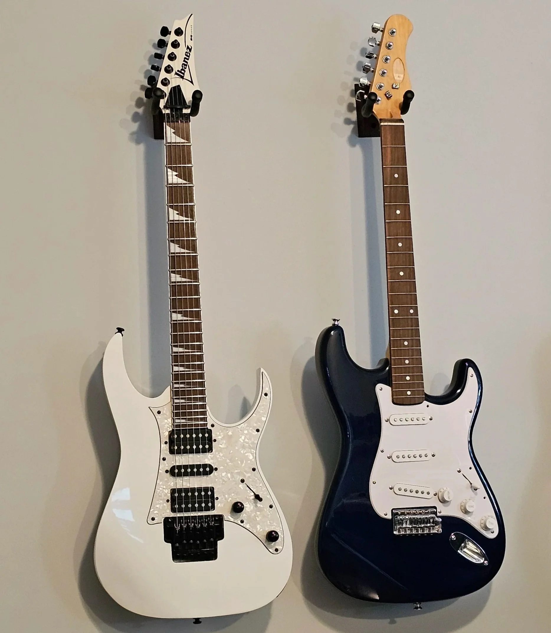 Two electric guitars hanging on a wall, one white with a patterned pickguard and the other black with a traditional pickguard.