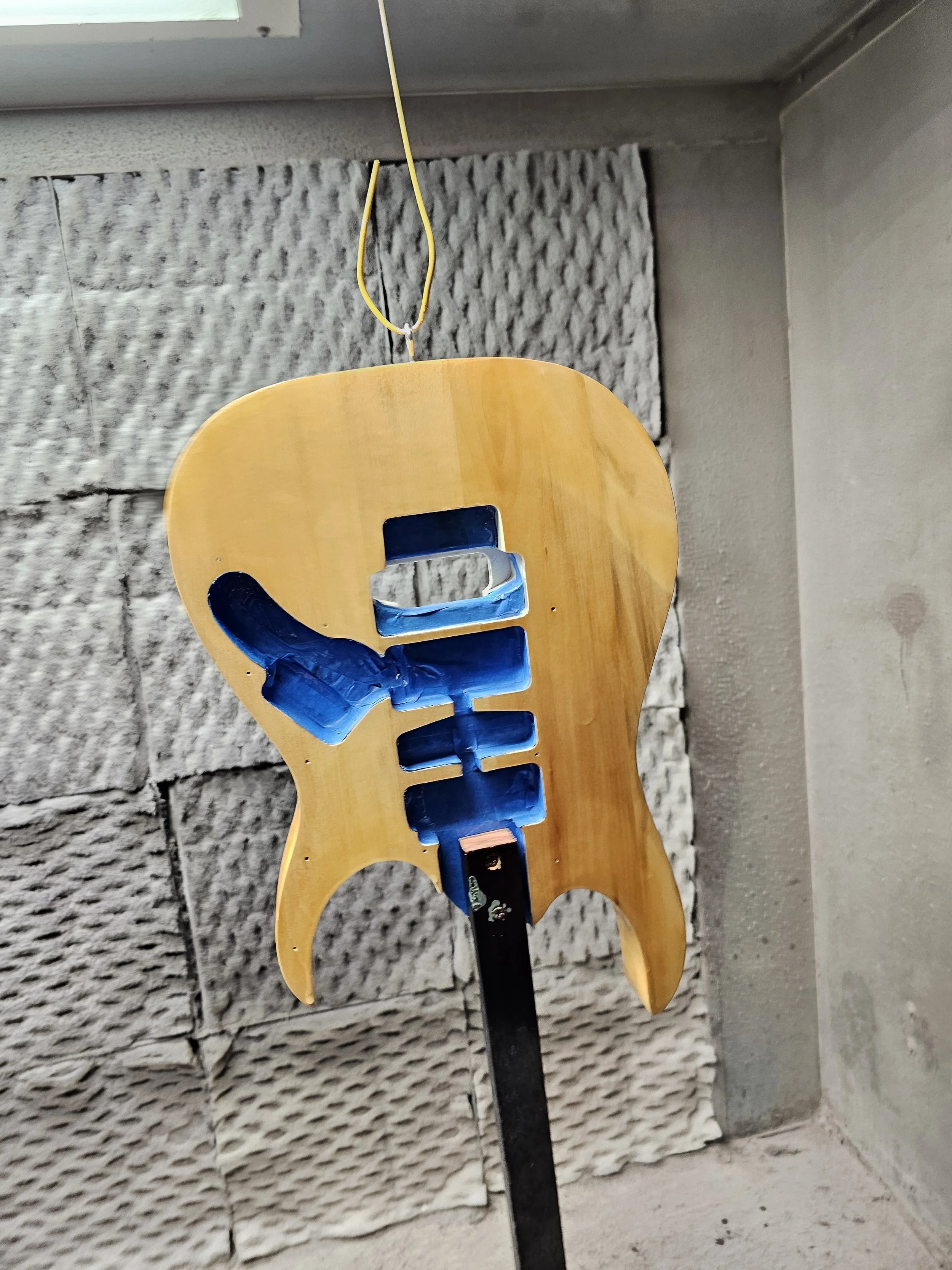A wooden guitar-shaped object, possibly a decoration or tool, mounted on a black stand with a backdrop of textured concrete blocks and a wall.