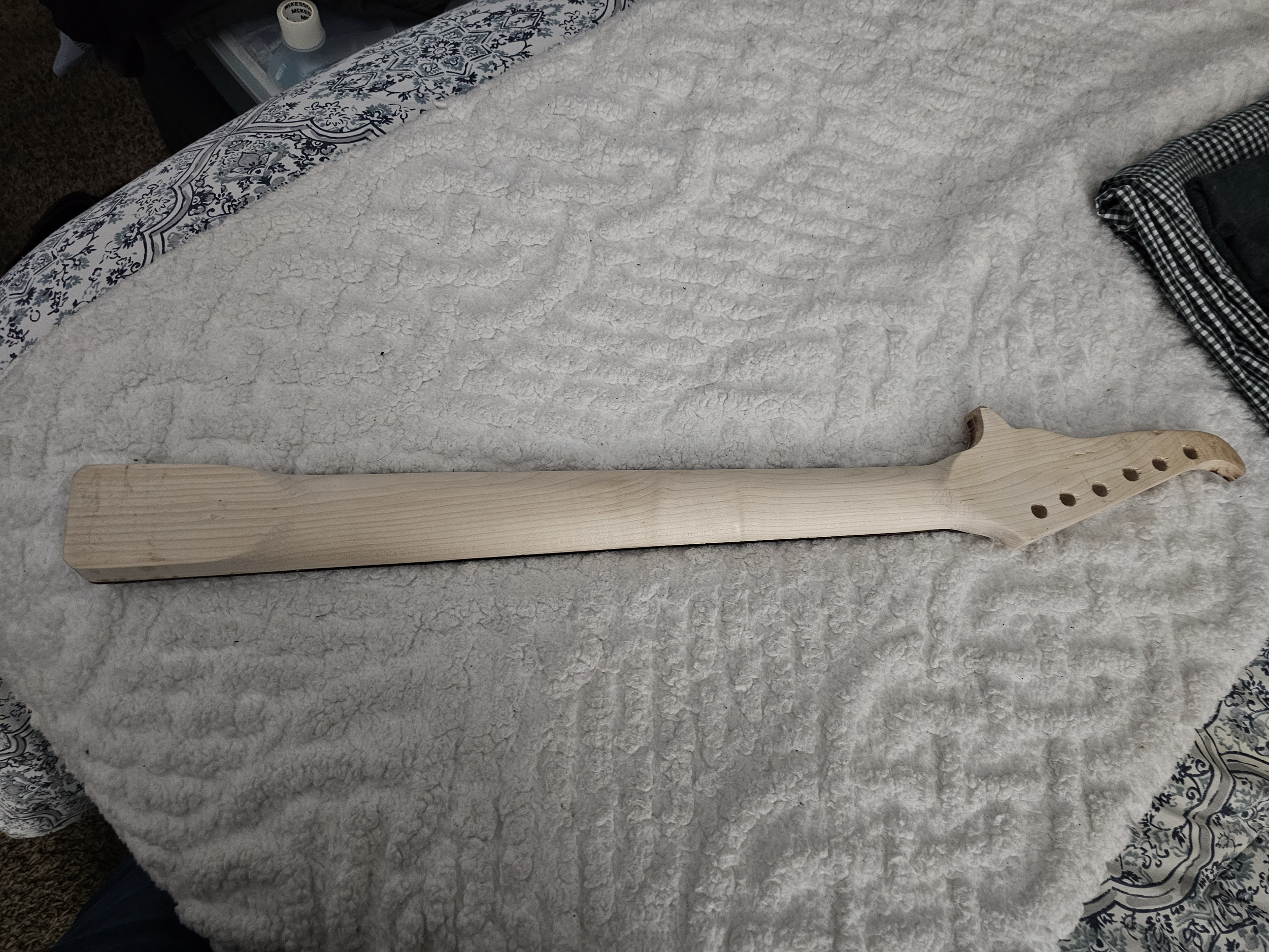 A wooden object with a flat rectangular end and a curved head with multiple holes, resembling a guitar neck or a similar musical instrument part, is lying on a textured, cream-colored surface.