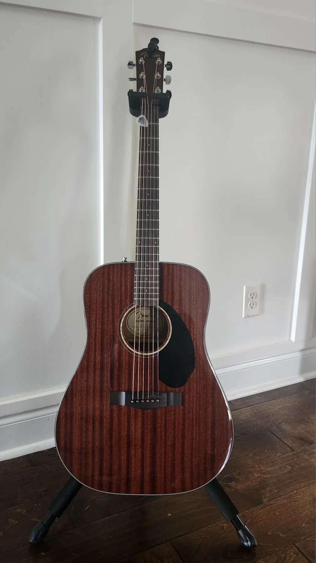 An acoustic guitar with a wooden finish standing on a black guitar stand on a hardwood floor next to a white wall and an electrical outlet.