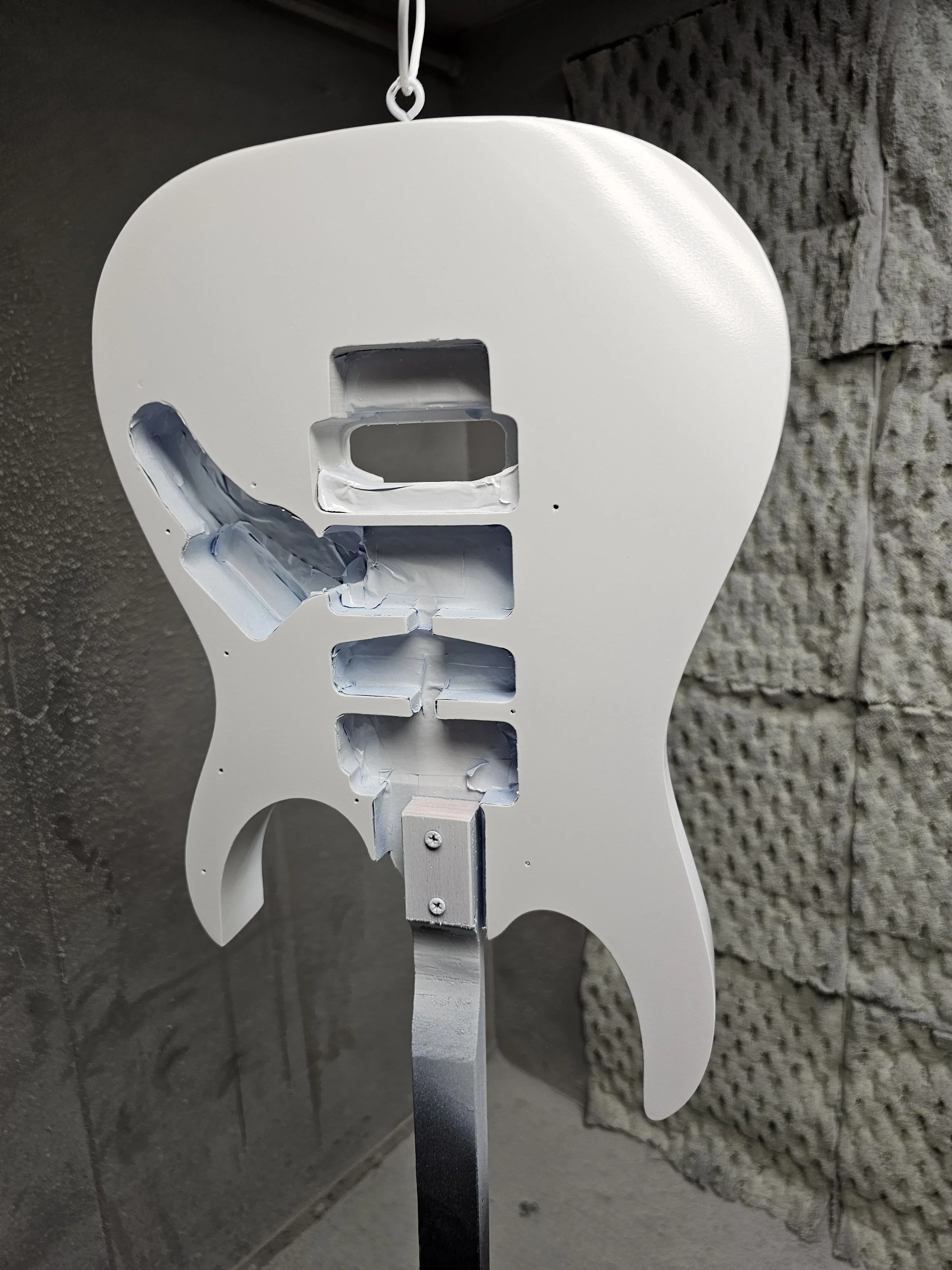Unpainted guitar body frame with cutouts, held up by a stand, against a textured wall background.