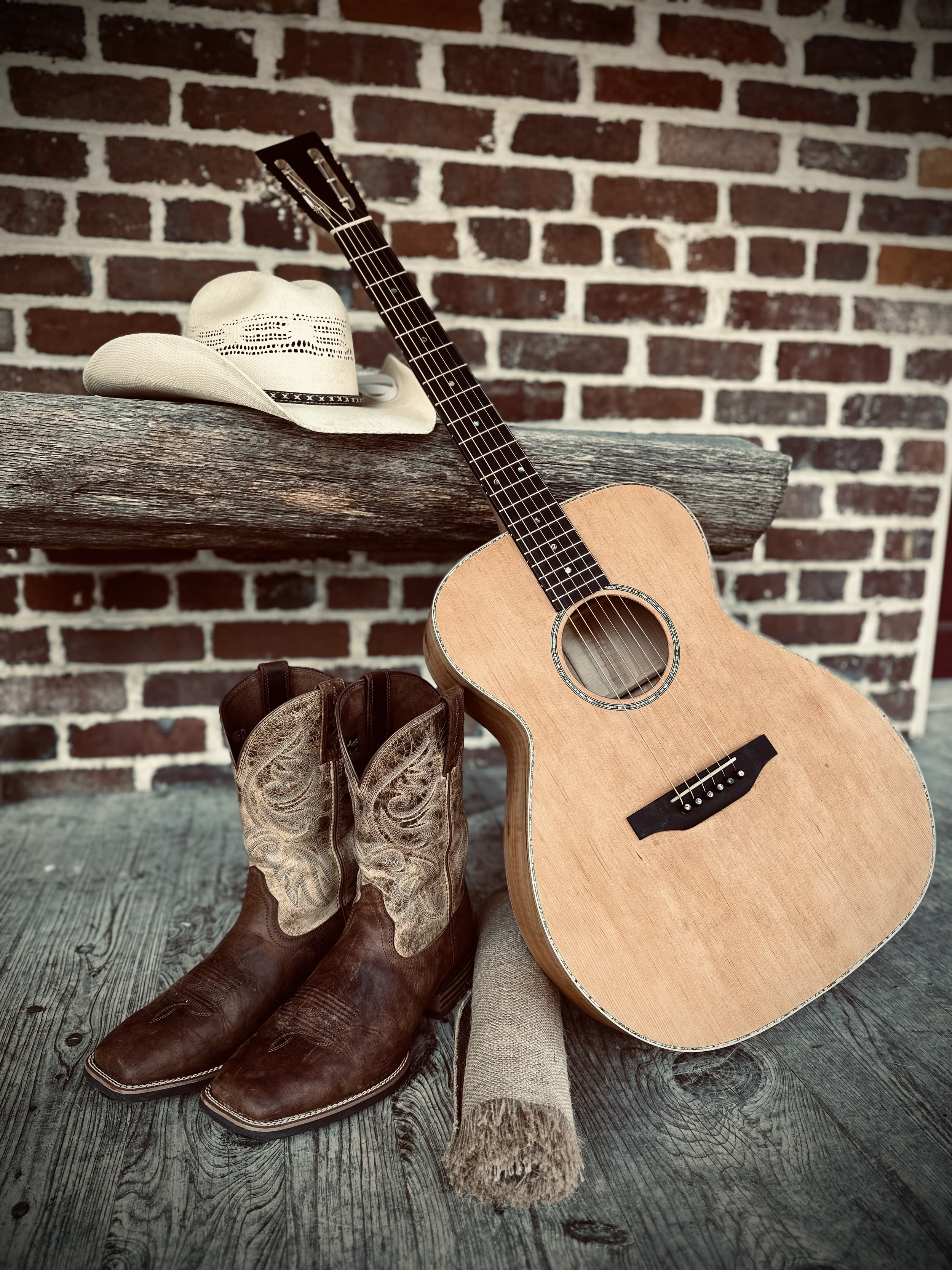 A cowboy hat, a guitar, cowboy boots, and a rolled-up rug on a wooden surface against a brick wall.
