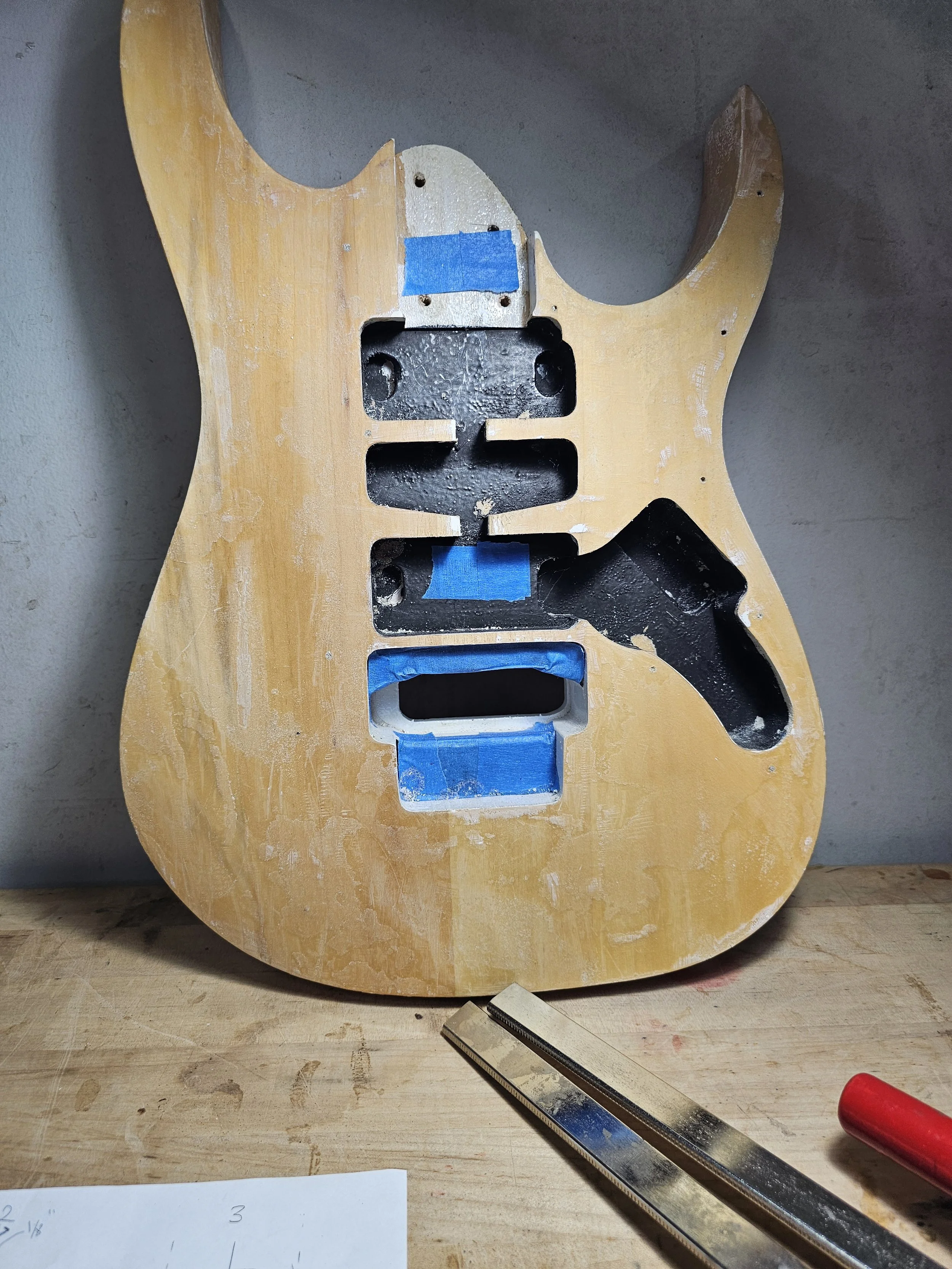 An unfinished electric guitar body made of wood, with cavities for pickups and electronics, taped off with blue painter's tape, positioned on a workbench with a metal ruler and part of a paper visible in the foreground.