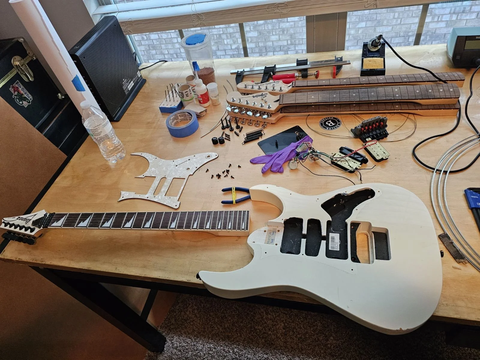 Disassembled electric guitar with a white body, neck, and headstock on a wooden table surrounded by guitar parts, tools, and electronic components.