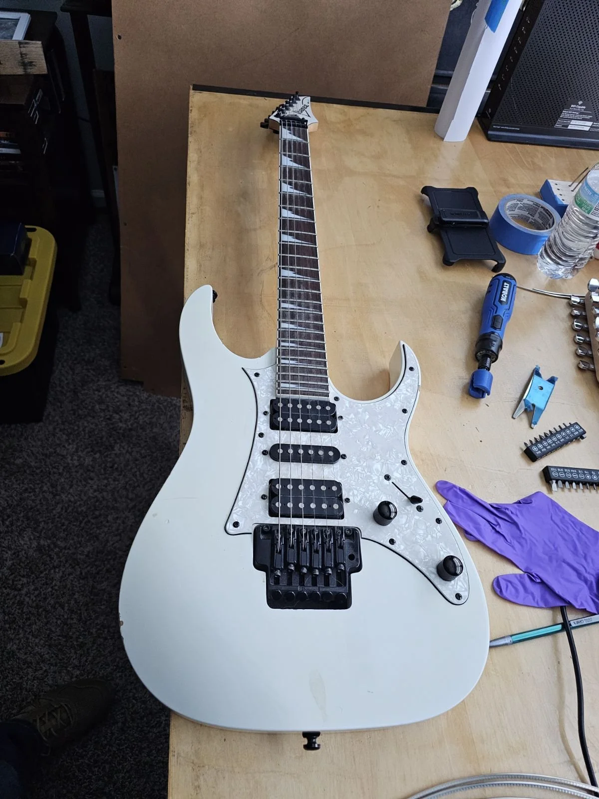 An electric guitar with a white body and black hardware resting on a workbench surrounded by tools and a purple glove.