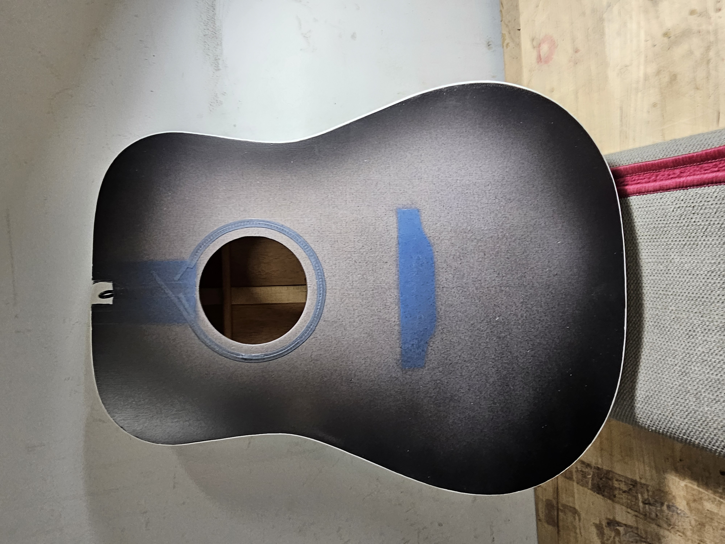 The back of an acoustic guitar, with the sound hole covered with blue painter's tape, resting against a wall and on top of a gray fabric surface.
