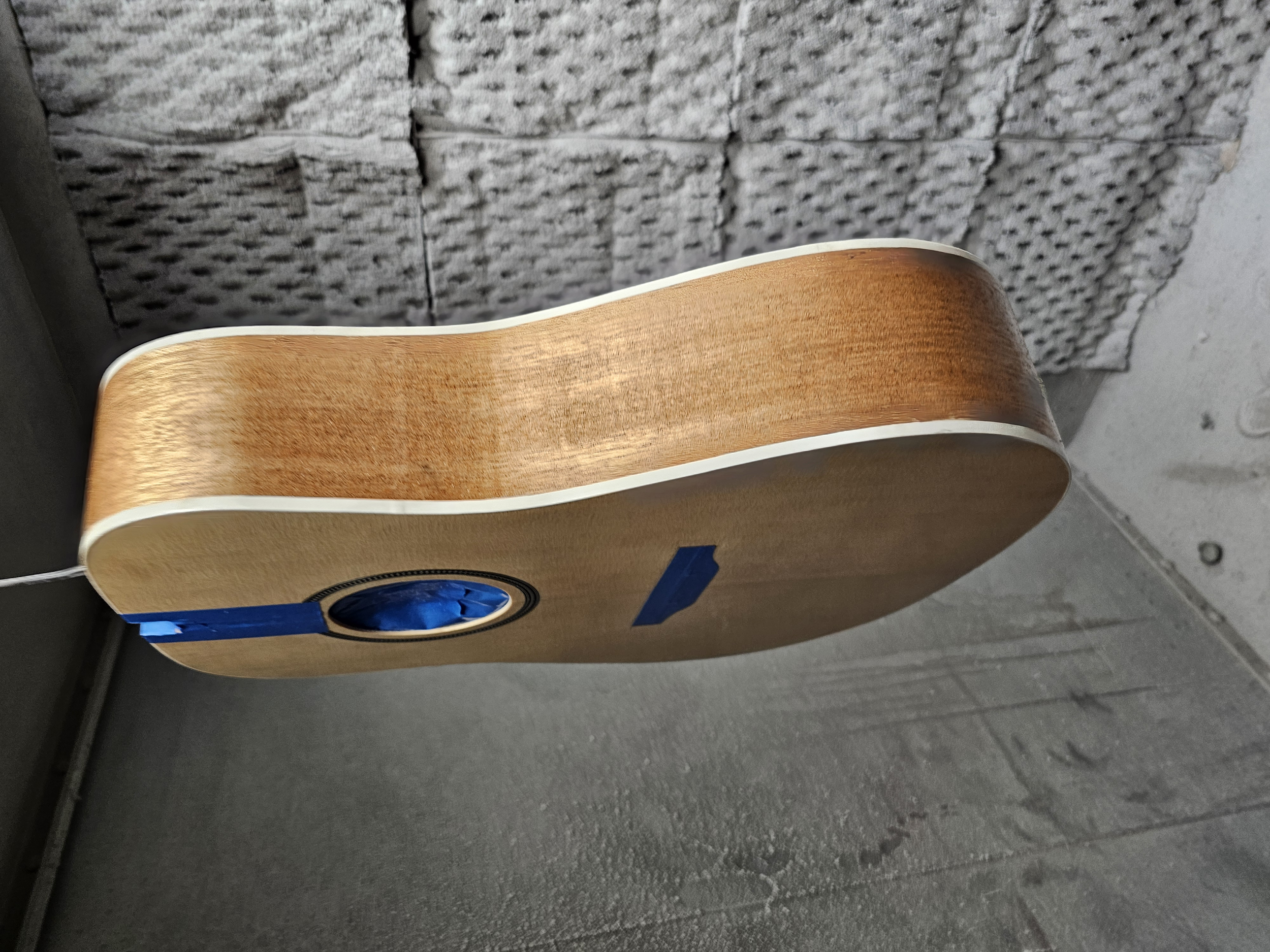 An acoustic guitar with a wooden body, lying against a rough stone wall, and partly covered with blue painter's tape.