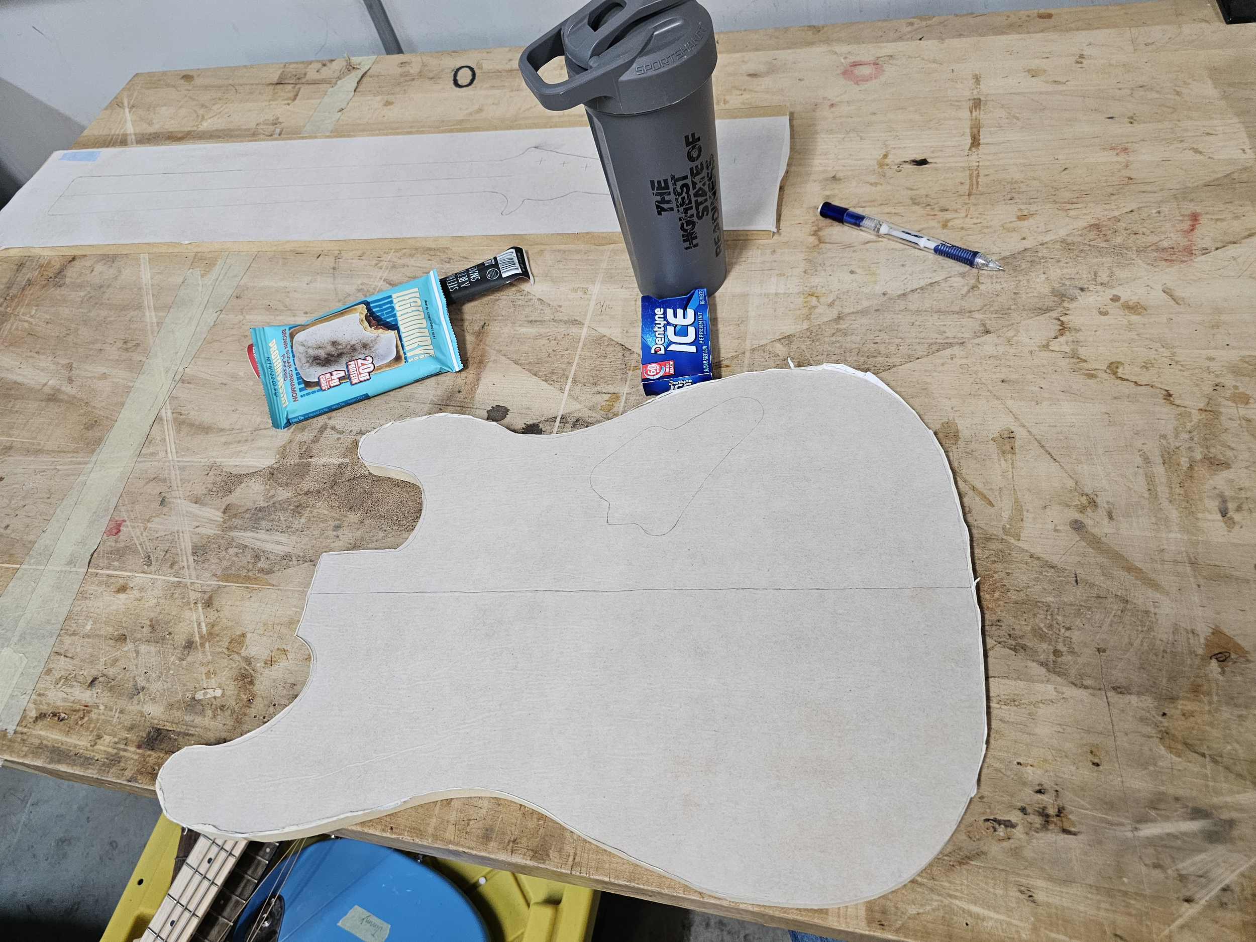 Unfinished bear-shaped cardboard cutout on a wooden workbench, surrounded by a tube of glue, a bag of miniature marshmallows, a container of ice, a blue pen, and a gray water bottle.