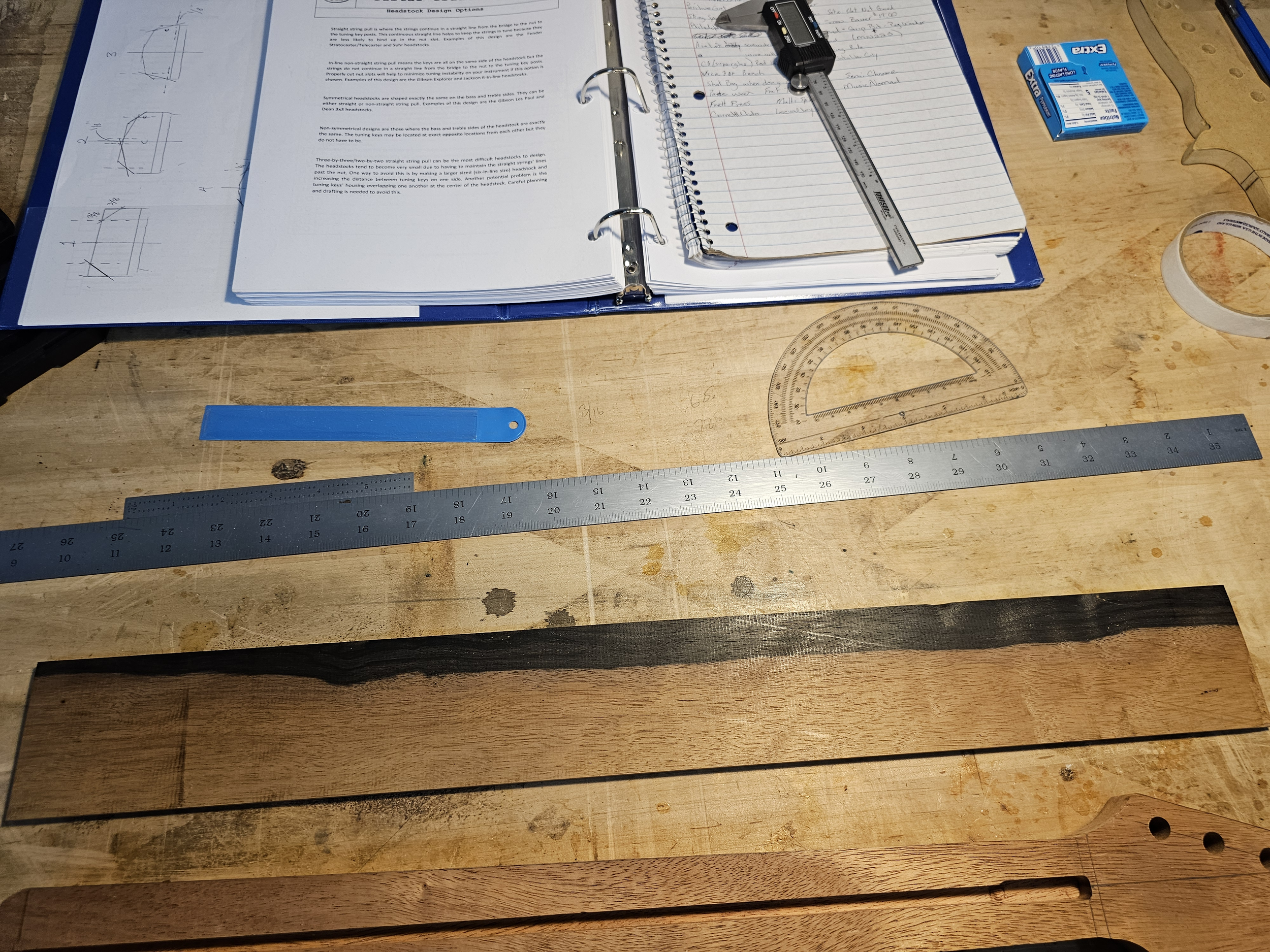 A woodworking workbench with various tools, including rulers, a protractor, a notepad, and sketches of a project, along with a piece of wood.