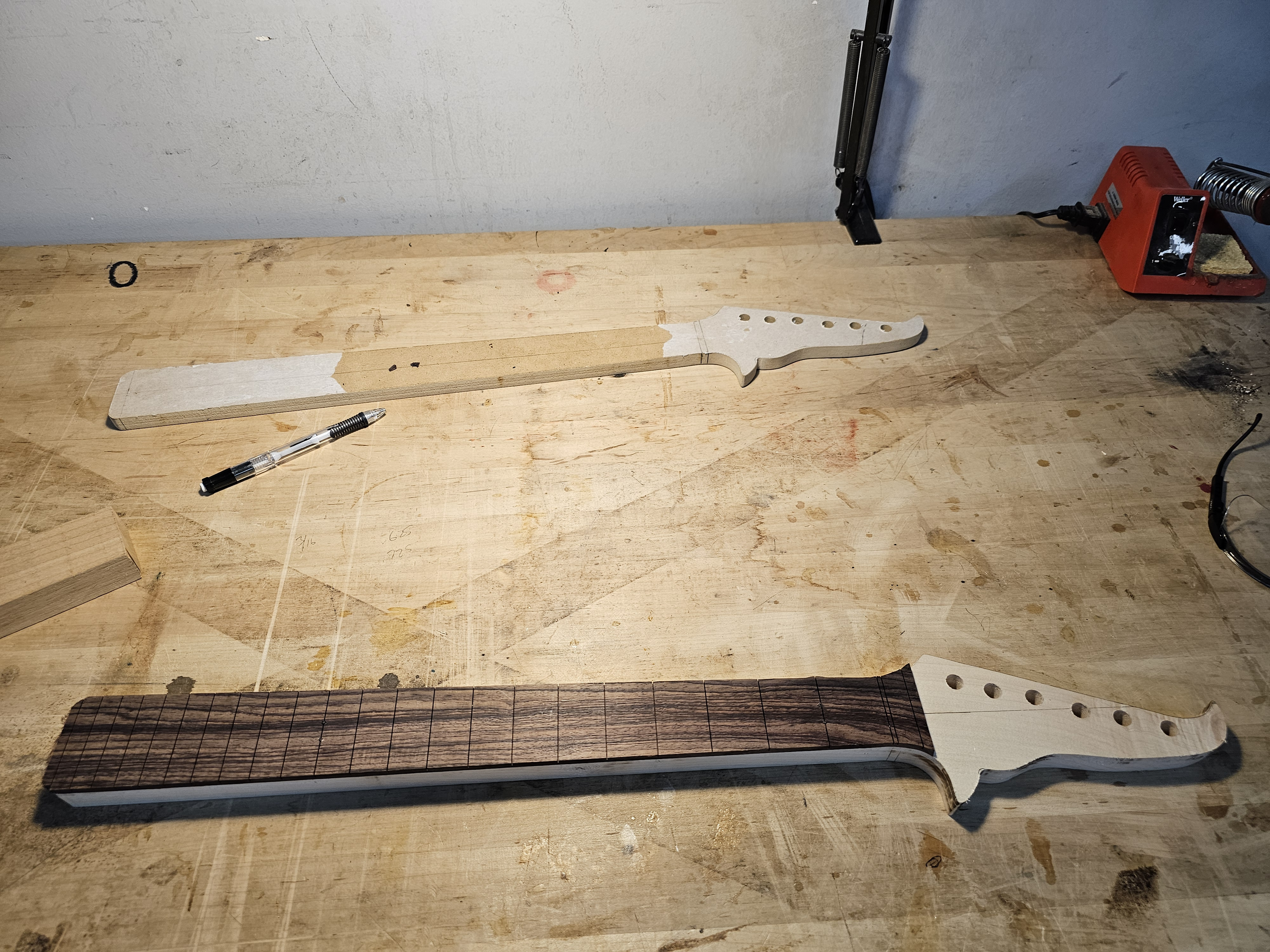 Two guitar necks in the process of construction on a woodworking table. One neck is partially painted white and the other is unpainted with a dark wood fretboard. Tools and parts, including a pen, a clamp, a soldering iron, and a small piece of wood,