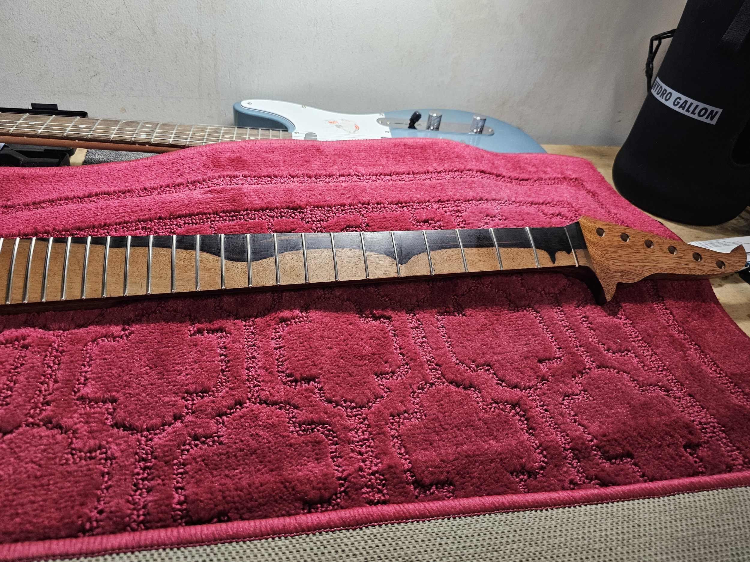 An electric guitar neck lying on a red towel on a table, with a pink towel beneath, and a black water gallon bottle and another much larger electric guitar in the background.