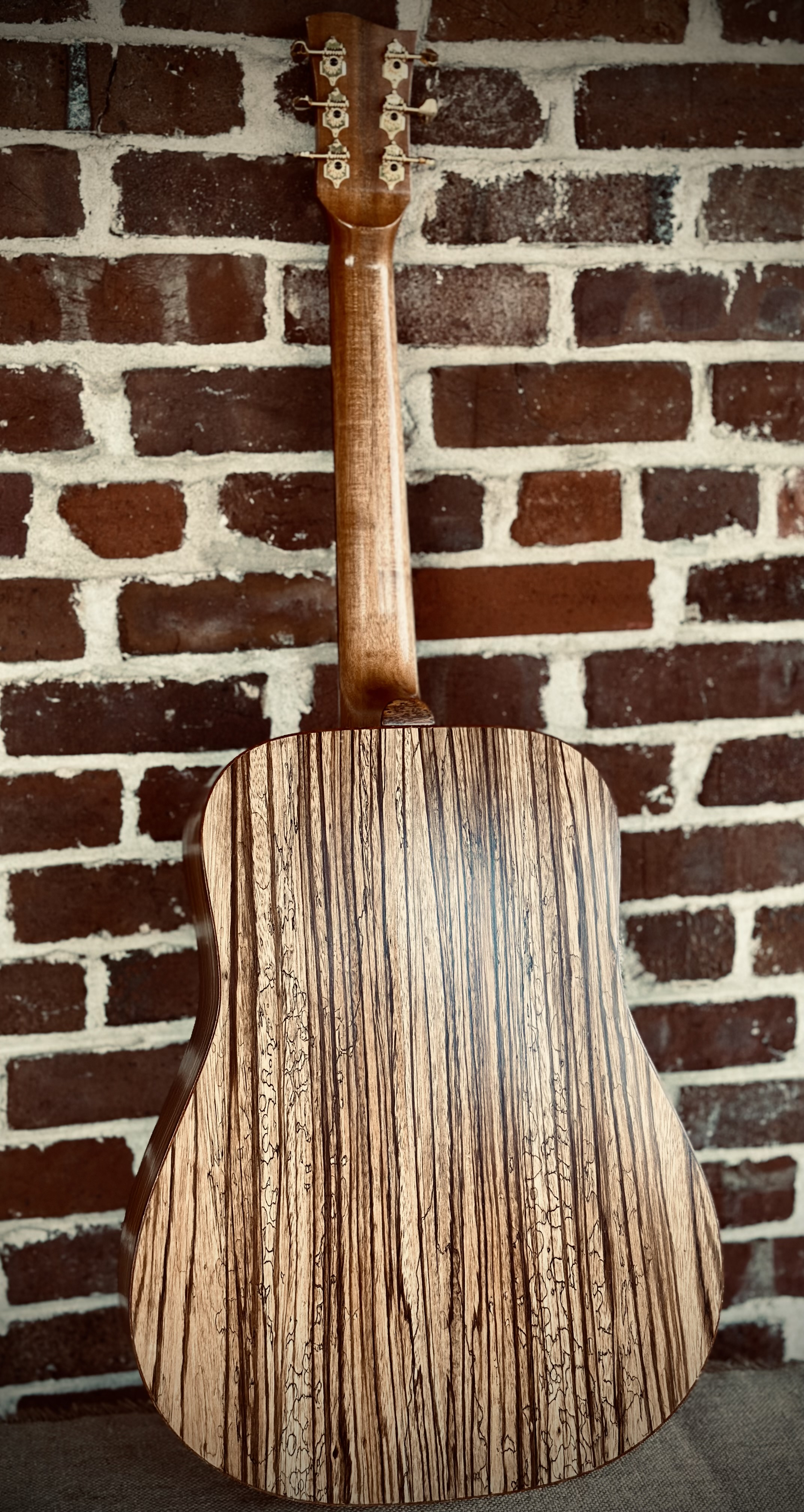 Back of a wooden acoustic guitar against a brick wall.