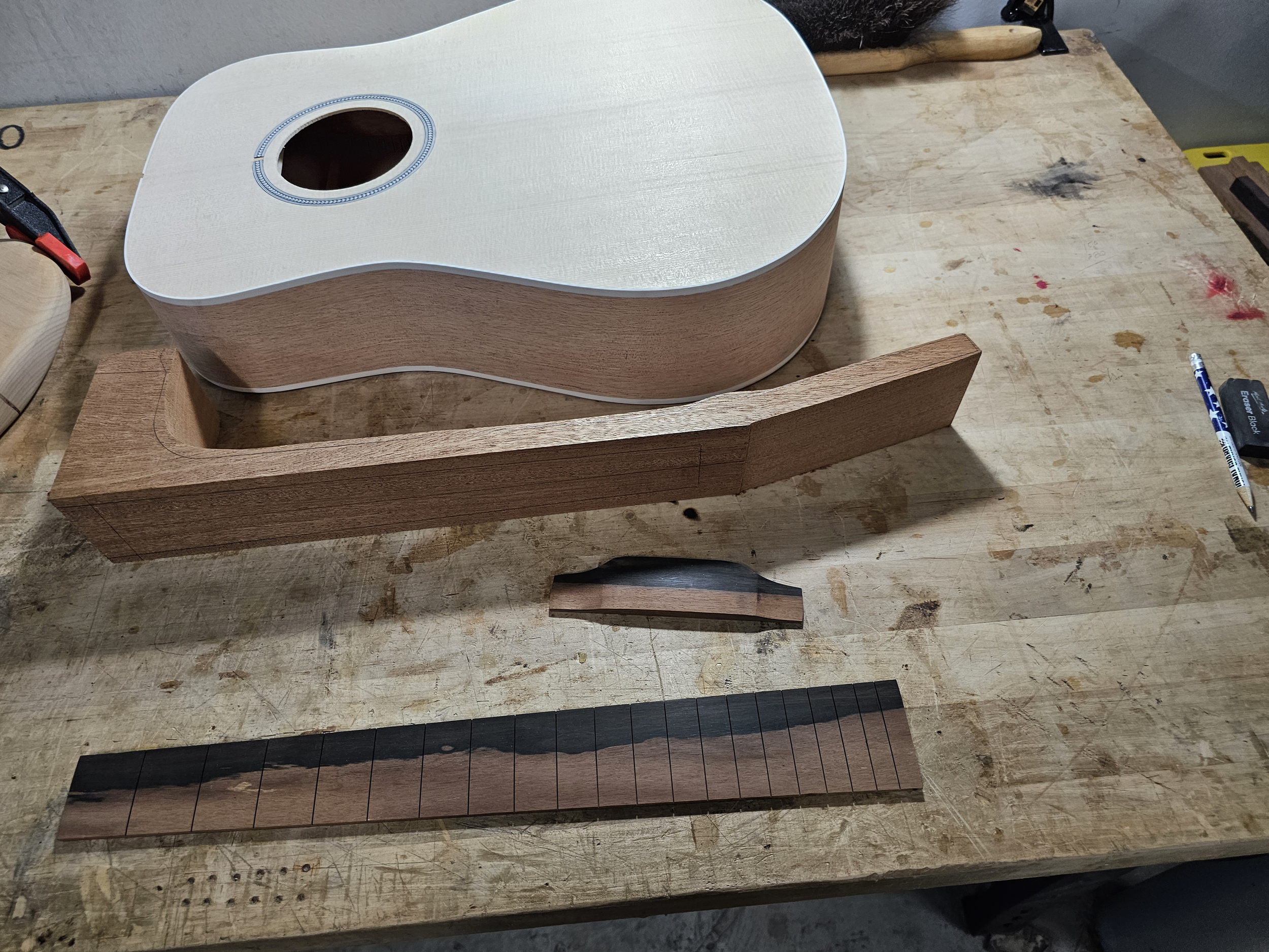 An unfinished wooden guitar body with a white finish and sound hole sitting on a workbench, alongside various pieces of wood and tools used for guitar construction.