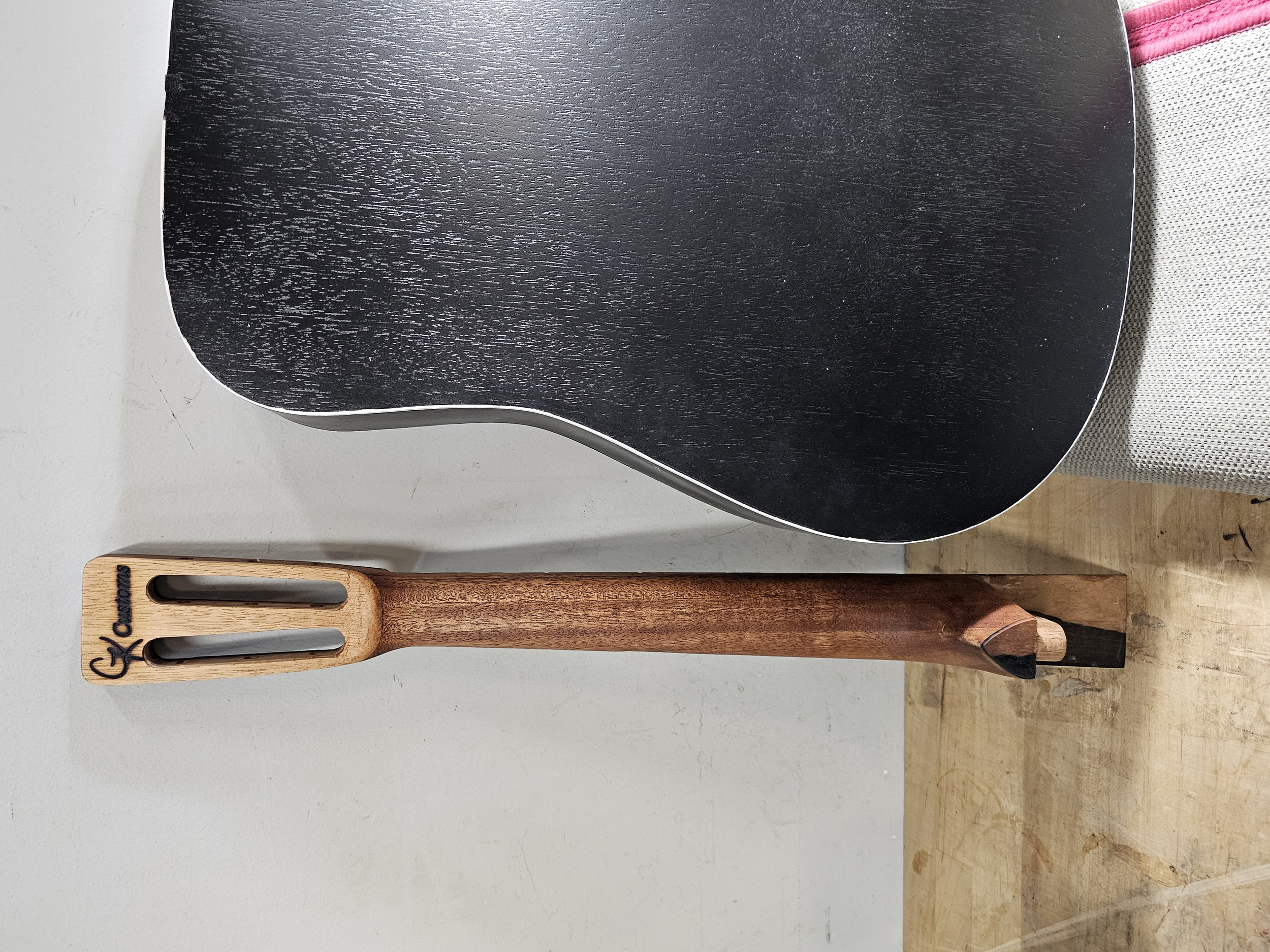 A black electric guitar with a curved body, placed on a white surface next to a wooden spatula with black slots and a branding logo.