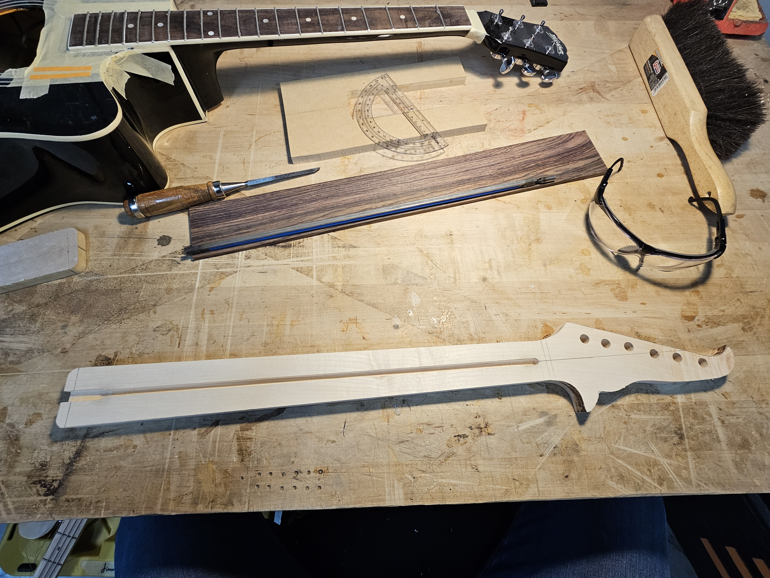 Wooden guitar neck template, various woodworking tools, glasses, and parts on a workbench.
