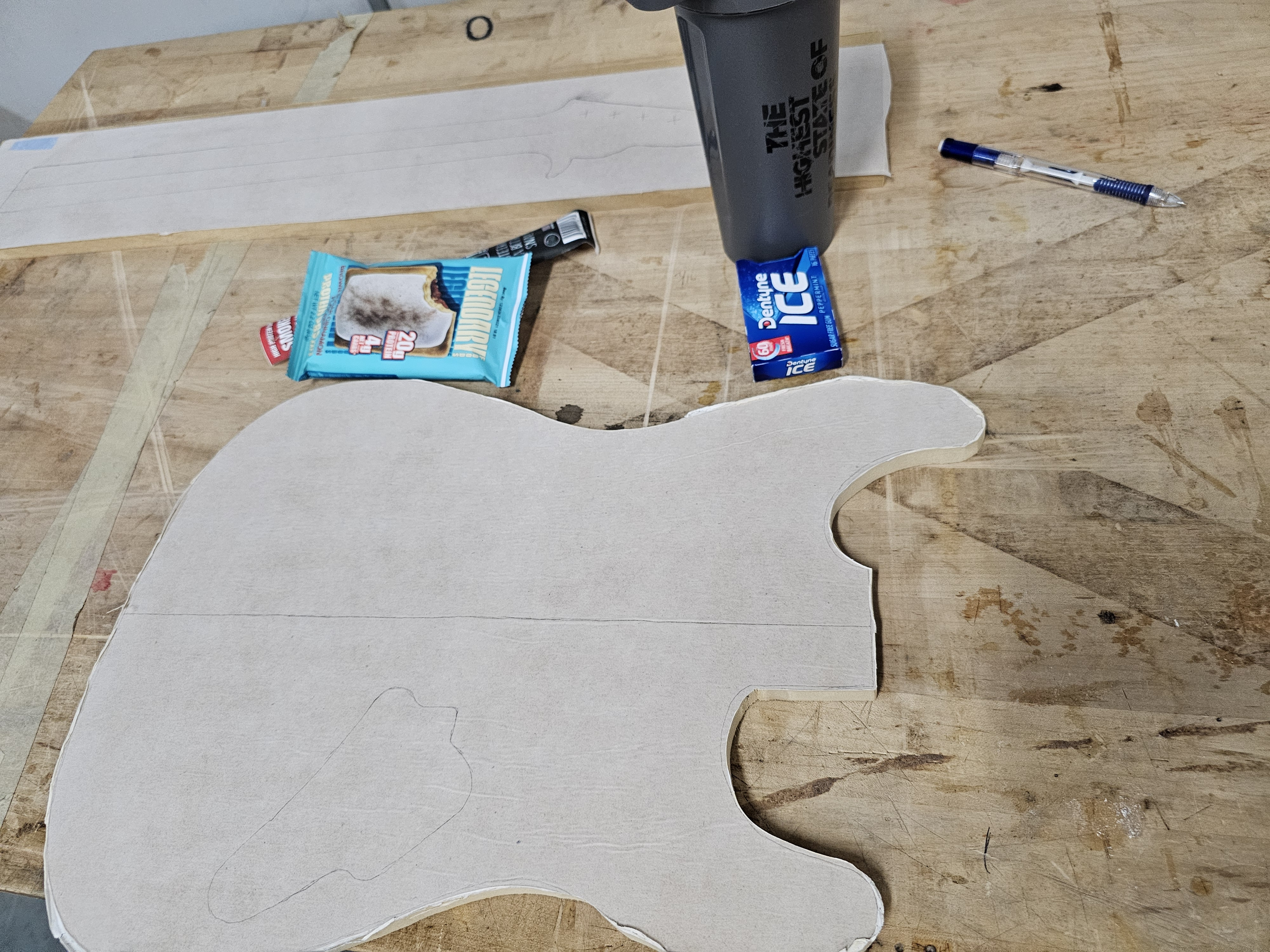 Un trabajo en progreso de una figura de cartón que se asemeja a un triceratops en una mesa de madera. Alrededor hay una barra de granola, un paquete de hielo, una taza con tapa, una pelota de pluma y una hoja de papel.