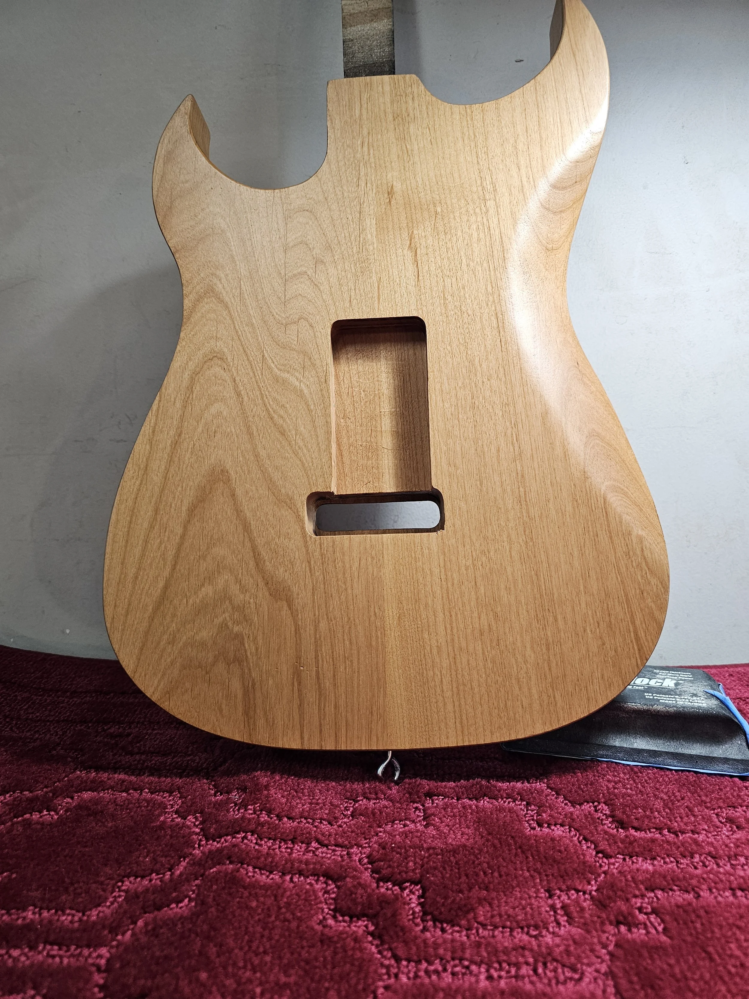 Unfinished wooden guitar body with cutouts, placed on a red textured fabric surface.