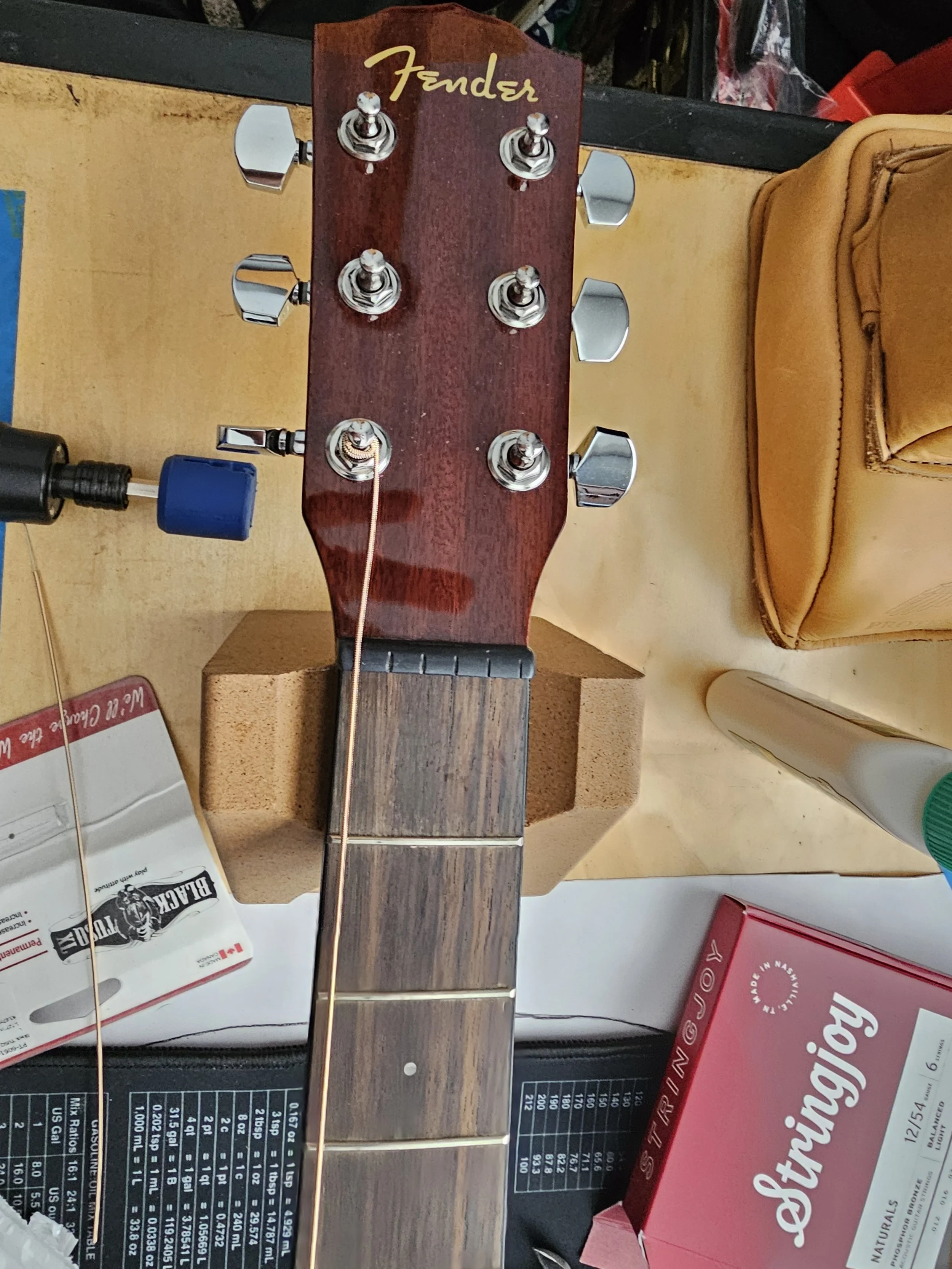 Close-up of a guitar headstock with six tuning pegs, labeled 'Fender', mounted on a workbench surrounded by tools and supplies.