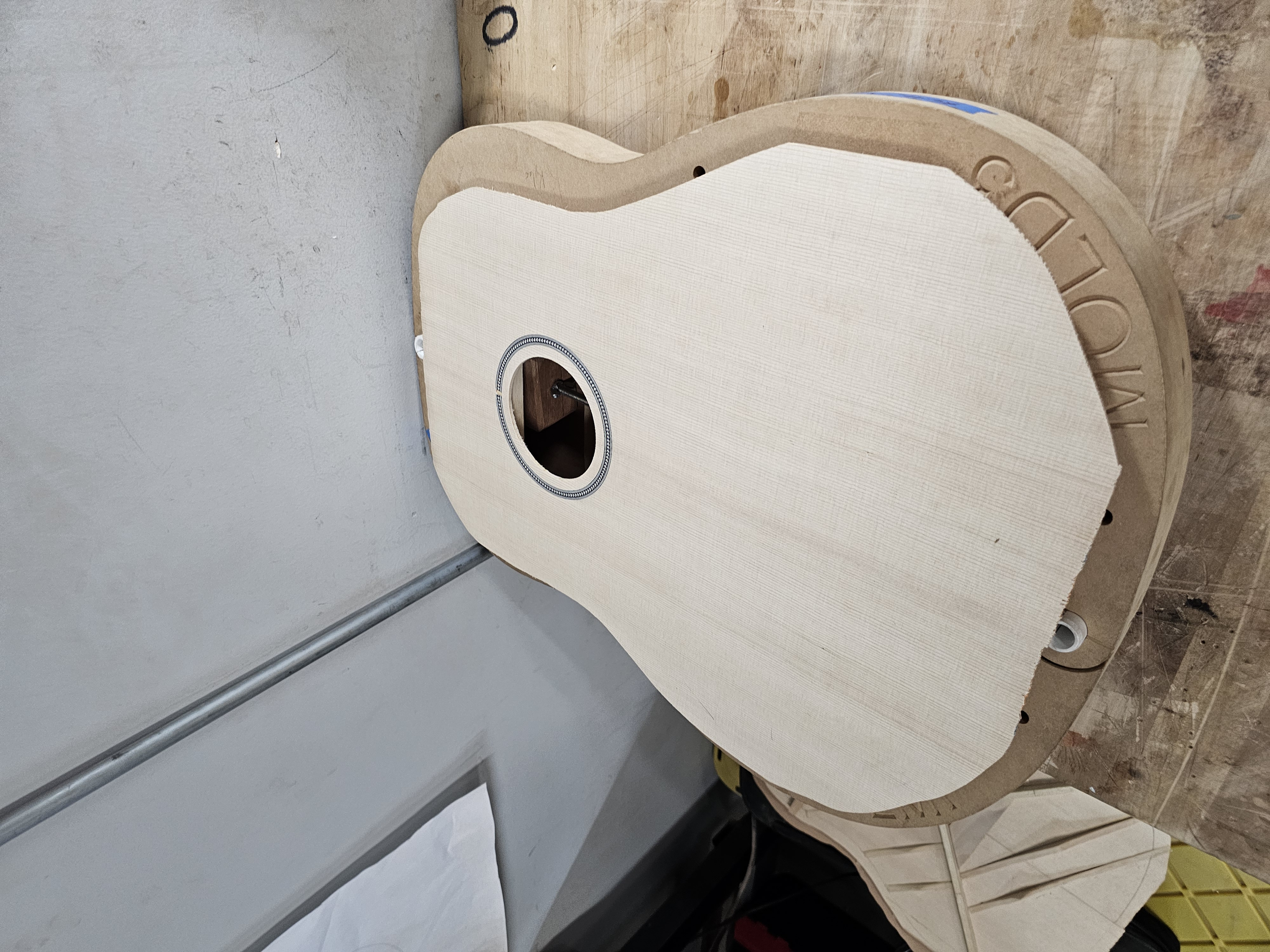 A guitar body under construction on a work table, with an opening for the soundhole and a partially attached beige fabric or padding.
