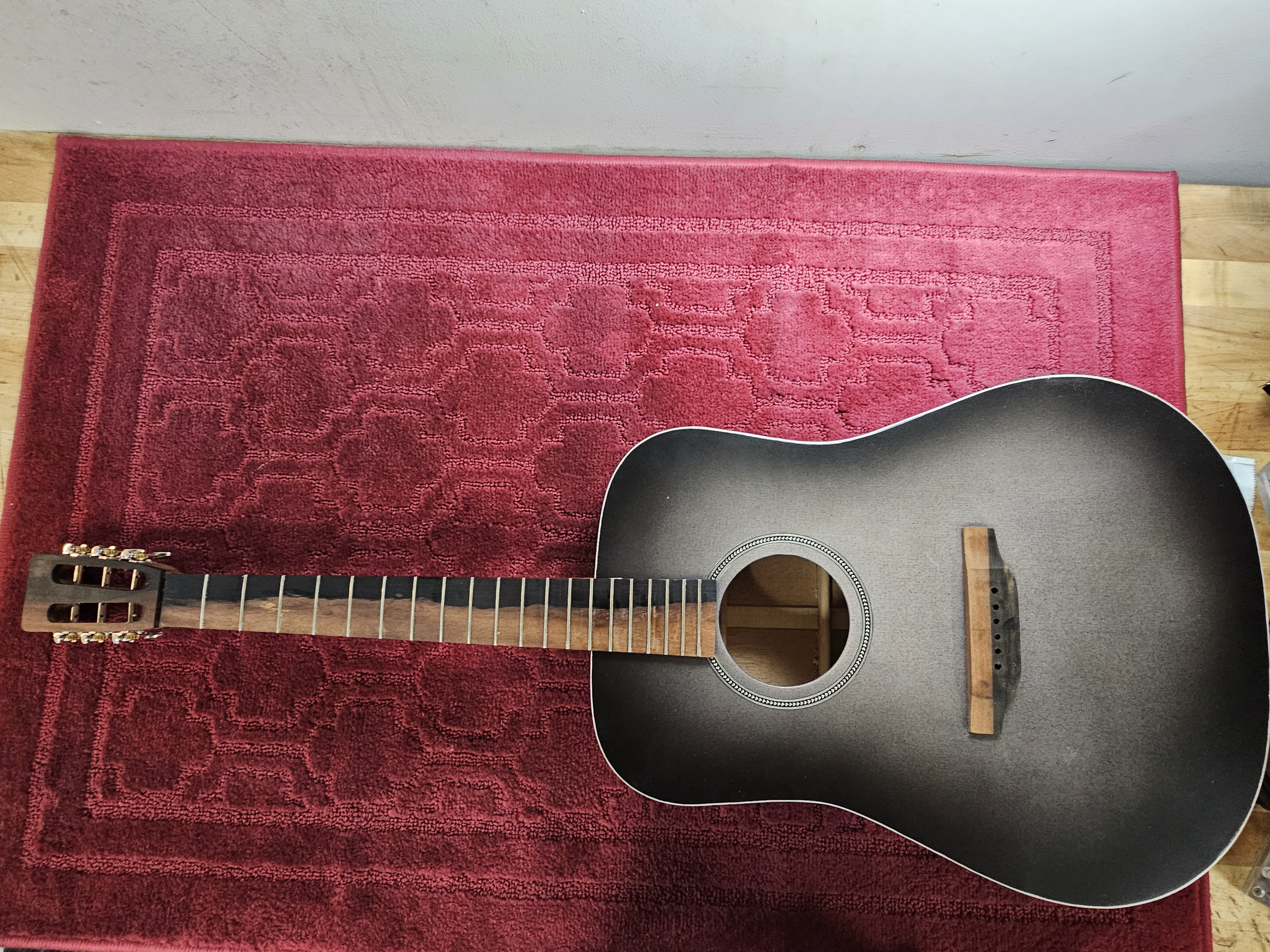 An acoustic guitar with a black body, no strings, and a light-colored wooden neck, resting on a red patterned rug on a wooden floor.