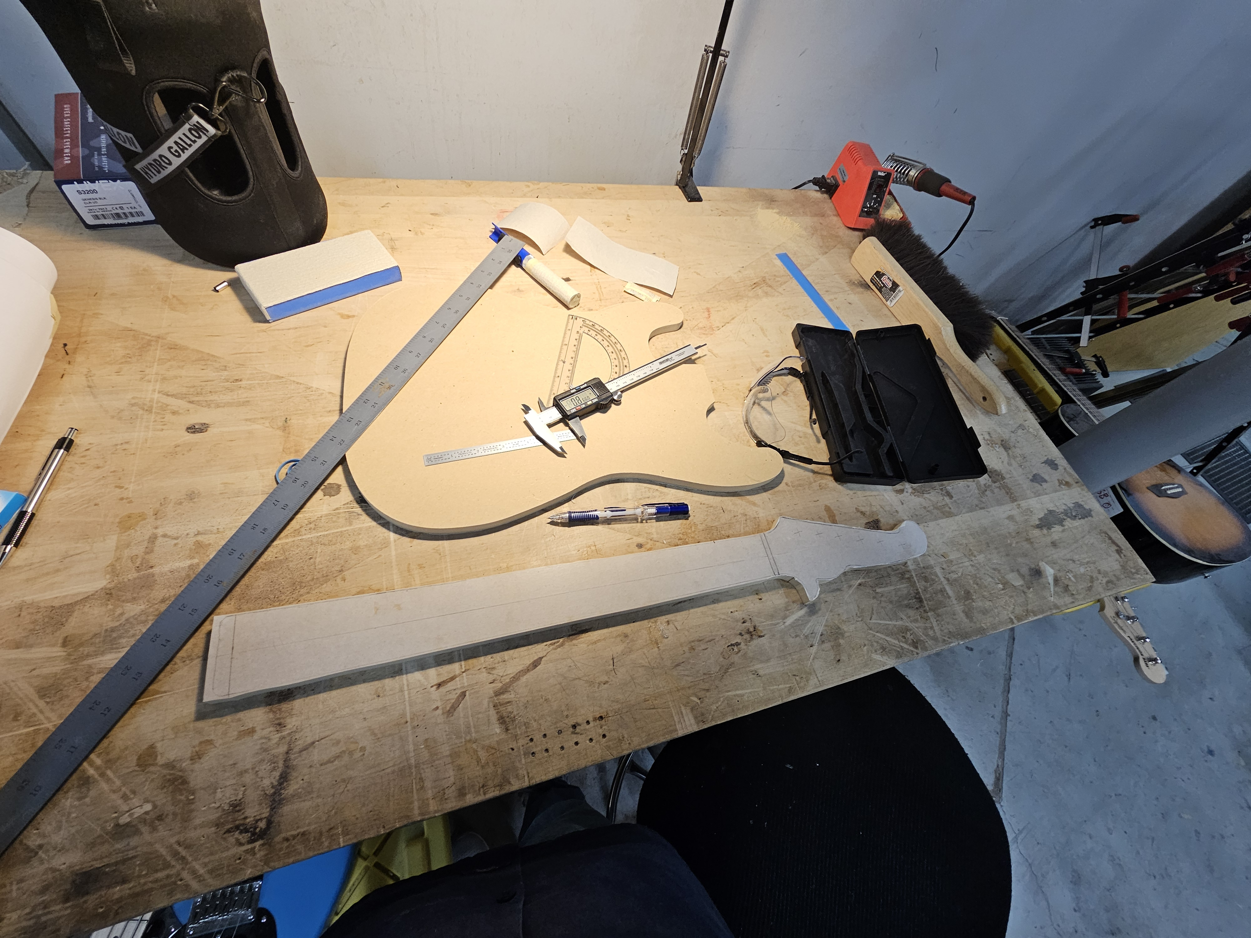 A workbench with woodworking tools and materials, including a guitar-shaped cutout, measuring ruler, caliper, pen, and templates, in a workshop setting.