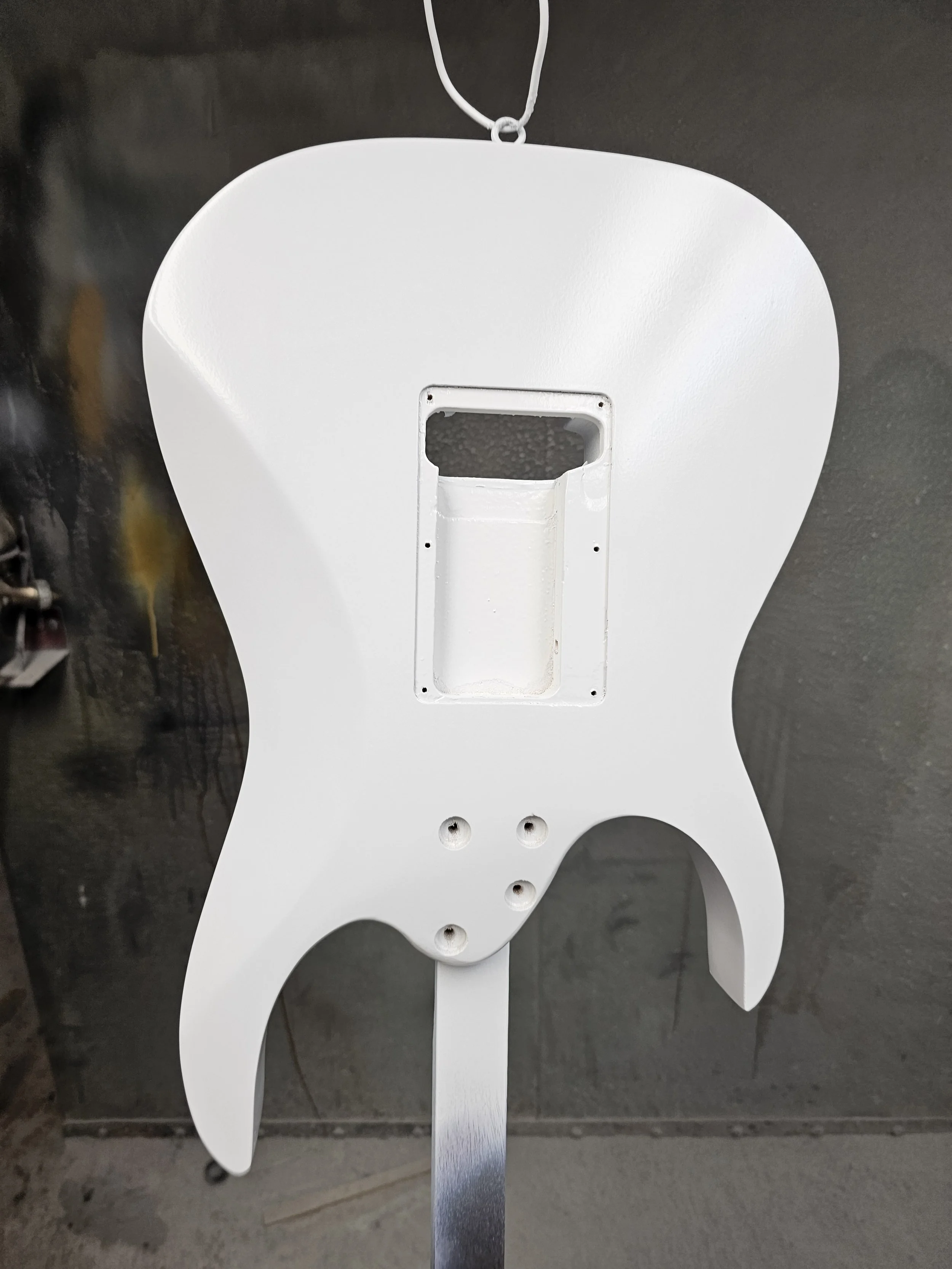 The image shows the unpainted body of an electric guitar, specifically the back side, with a white finish and visible screw holes for hardware. The guitar is hanging on a hook against a dark wall.