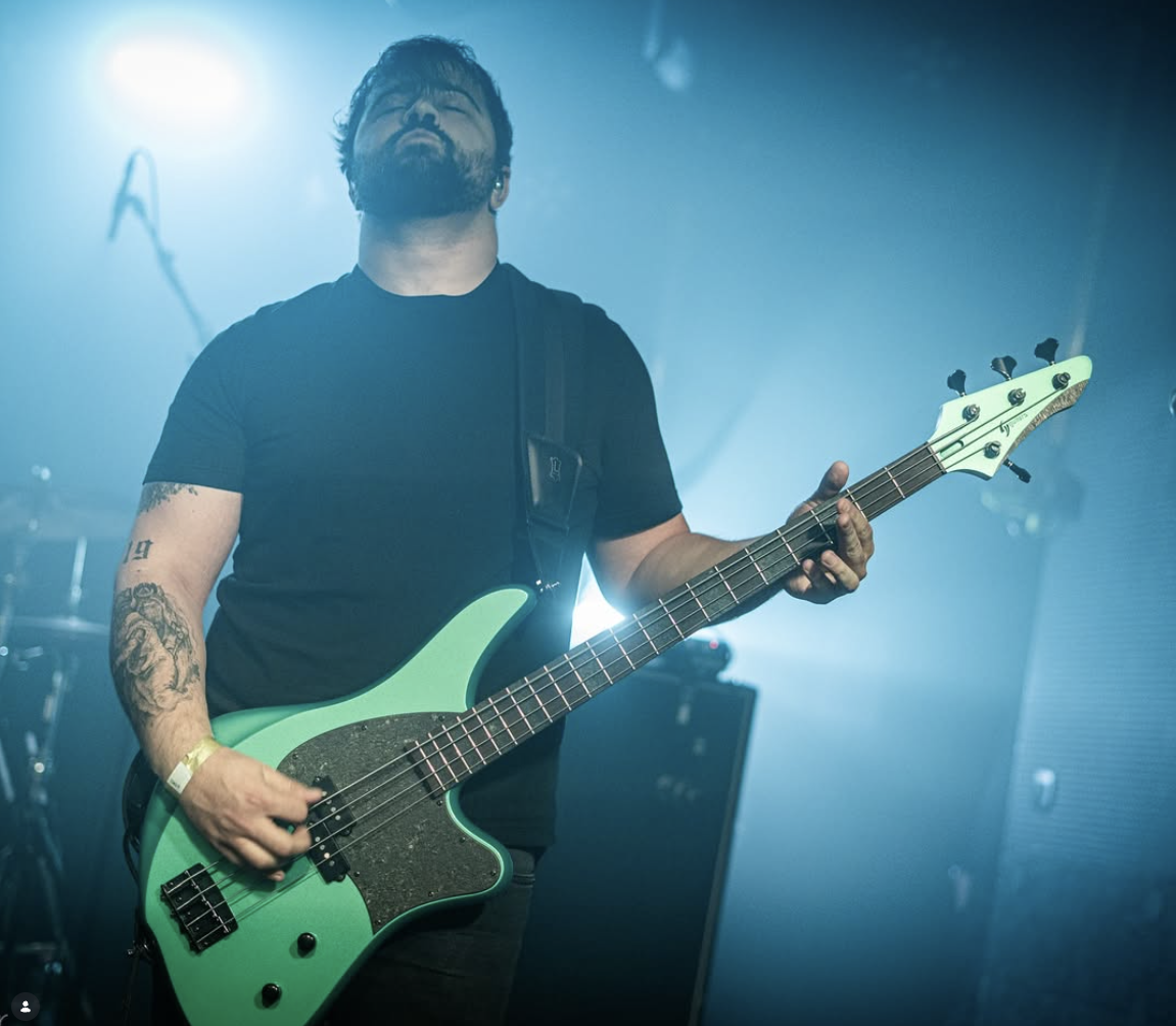 Bearded musician playing a bright green electric bass guitar on a blue-lit concert stage