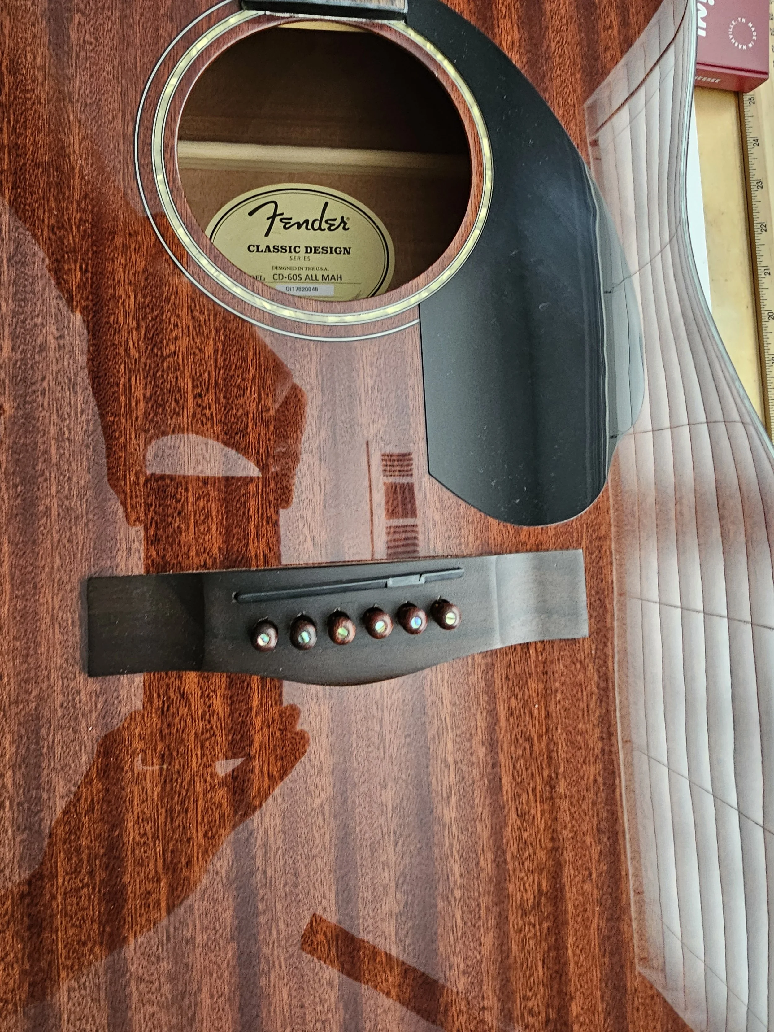 An acoustic guitar with a glossy wood finish, showing the sound hole and bridge, resting on a wooden surface. The inside label of the guitar indicates it is a Fender Classic Design series.