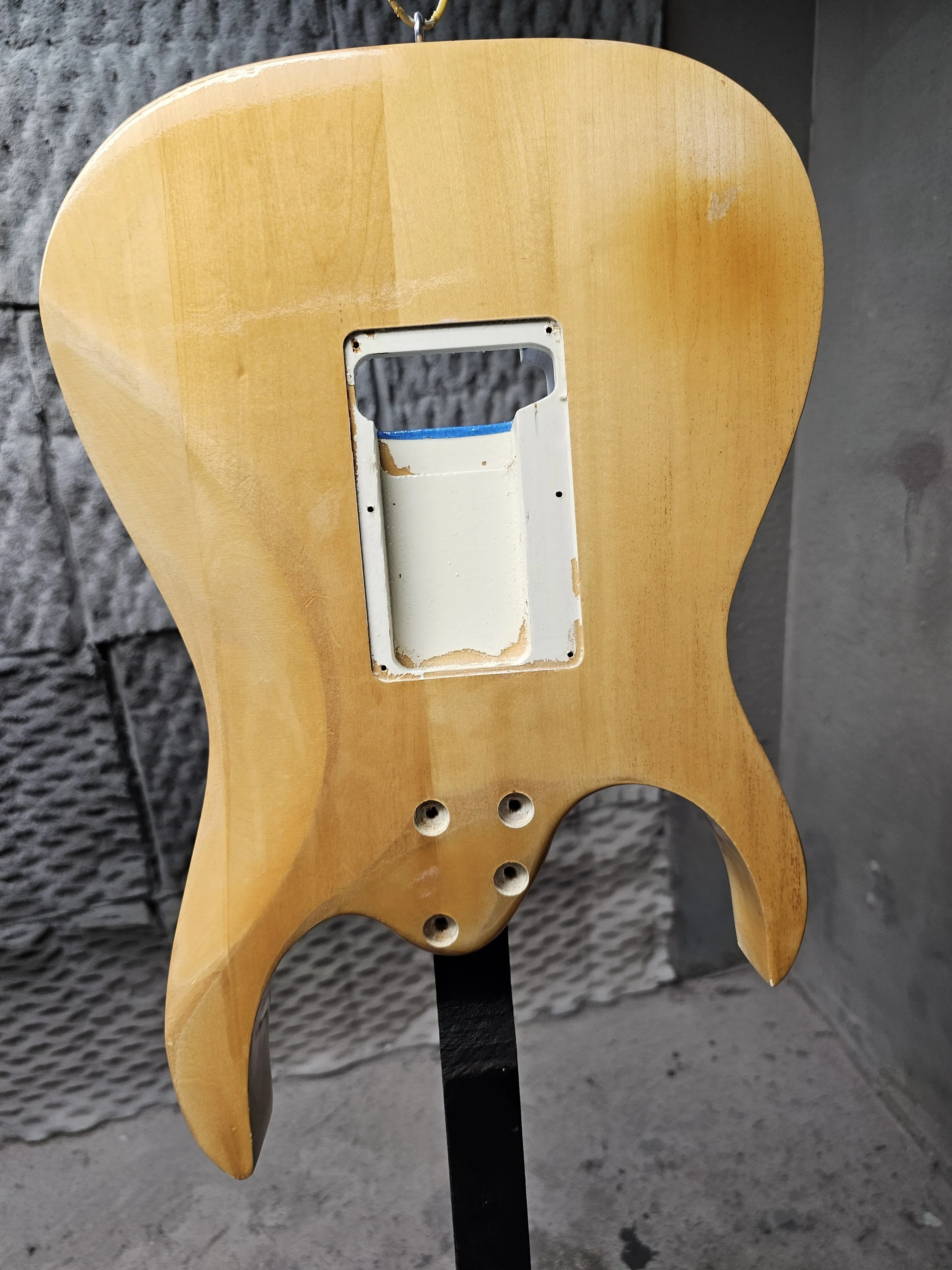 Wooden guitar body without pickups or electronics, mounted on a black stand, with a gray wall background.