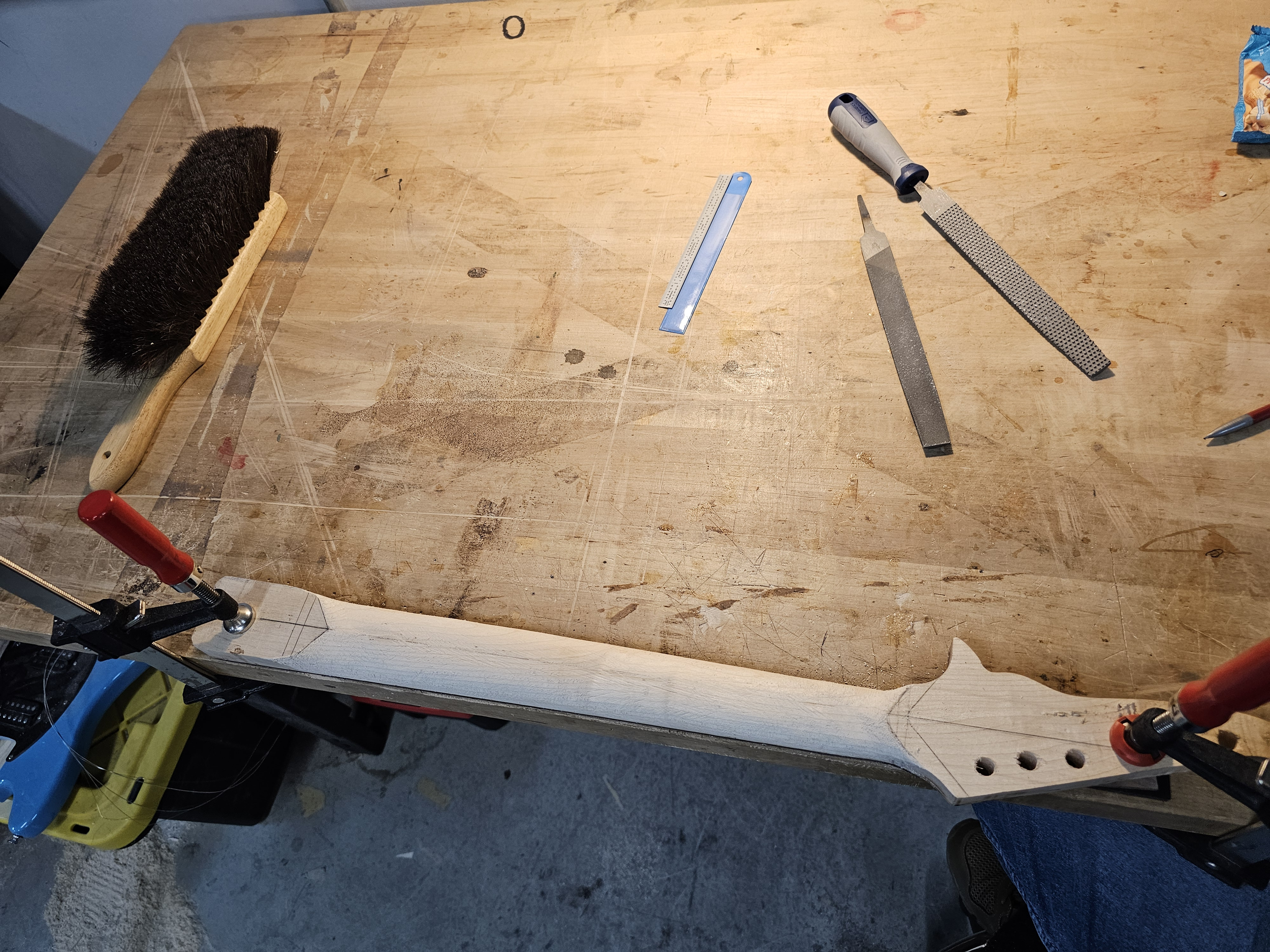 A woodworking workbench with tools including a brush, a square, a rasp, a file, and a chisel, with a piece of wood clamped to the edge.