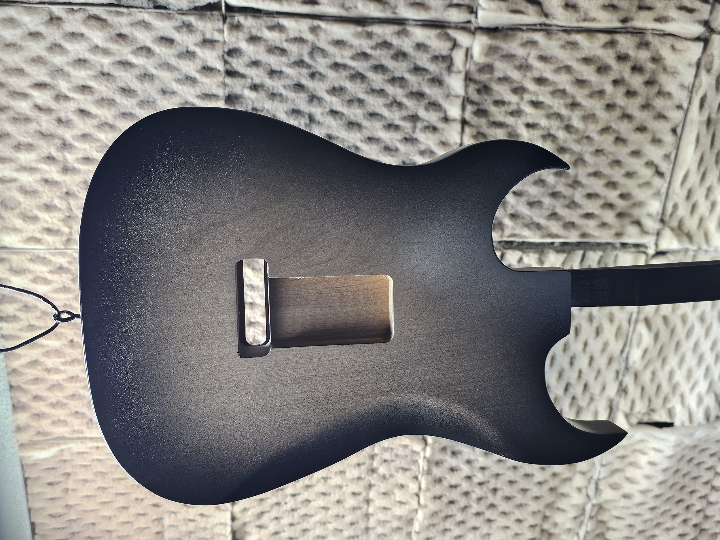 Close-up of a black electric guitar body with a rectangular hole in the center, against a textured brick wall background.