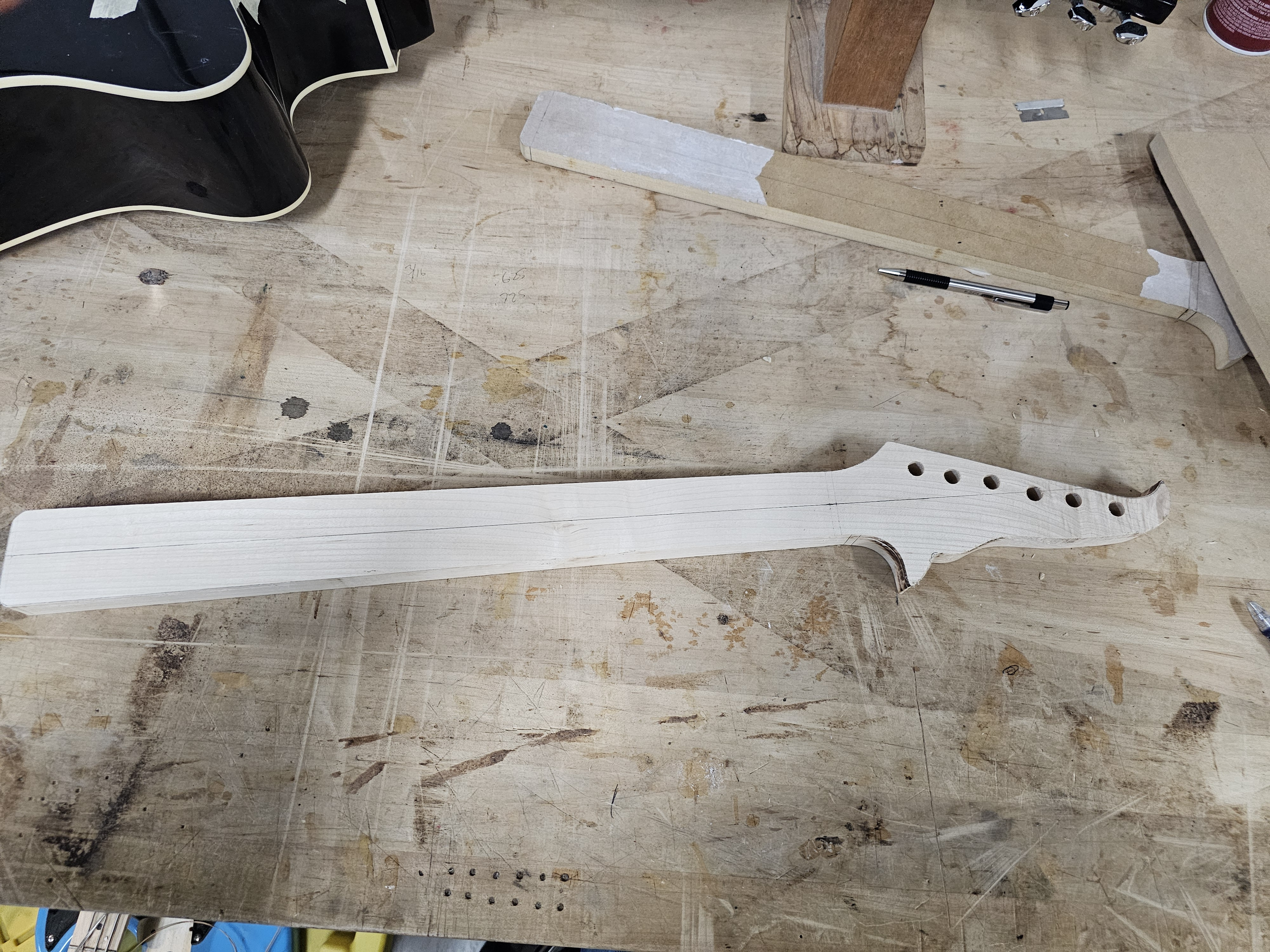 Unfinished wooden guitar neck on a workbench with various tools and wood pieces around.