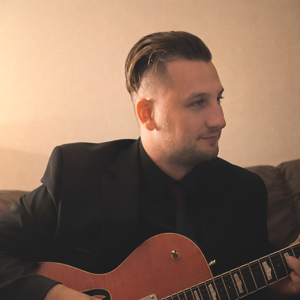 A man dressed in a black suit playing an acoustic guitar in a dimly lit room.