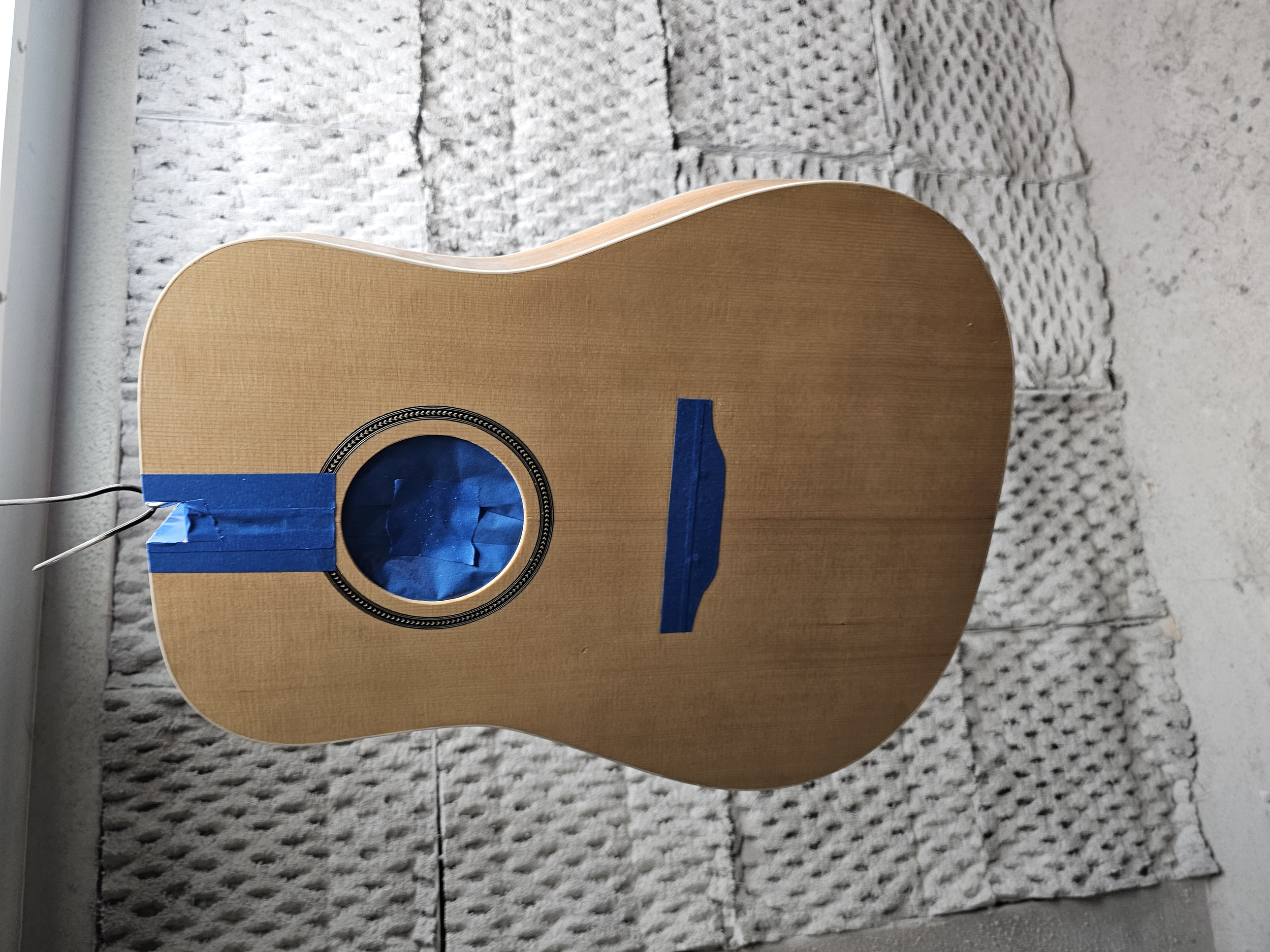 An unfinished acoustic guitar with masking tape covering the sound hole and a blue paper circle inside, placed on a carpeted floor against a brick wall.