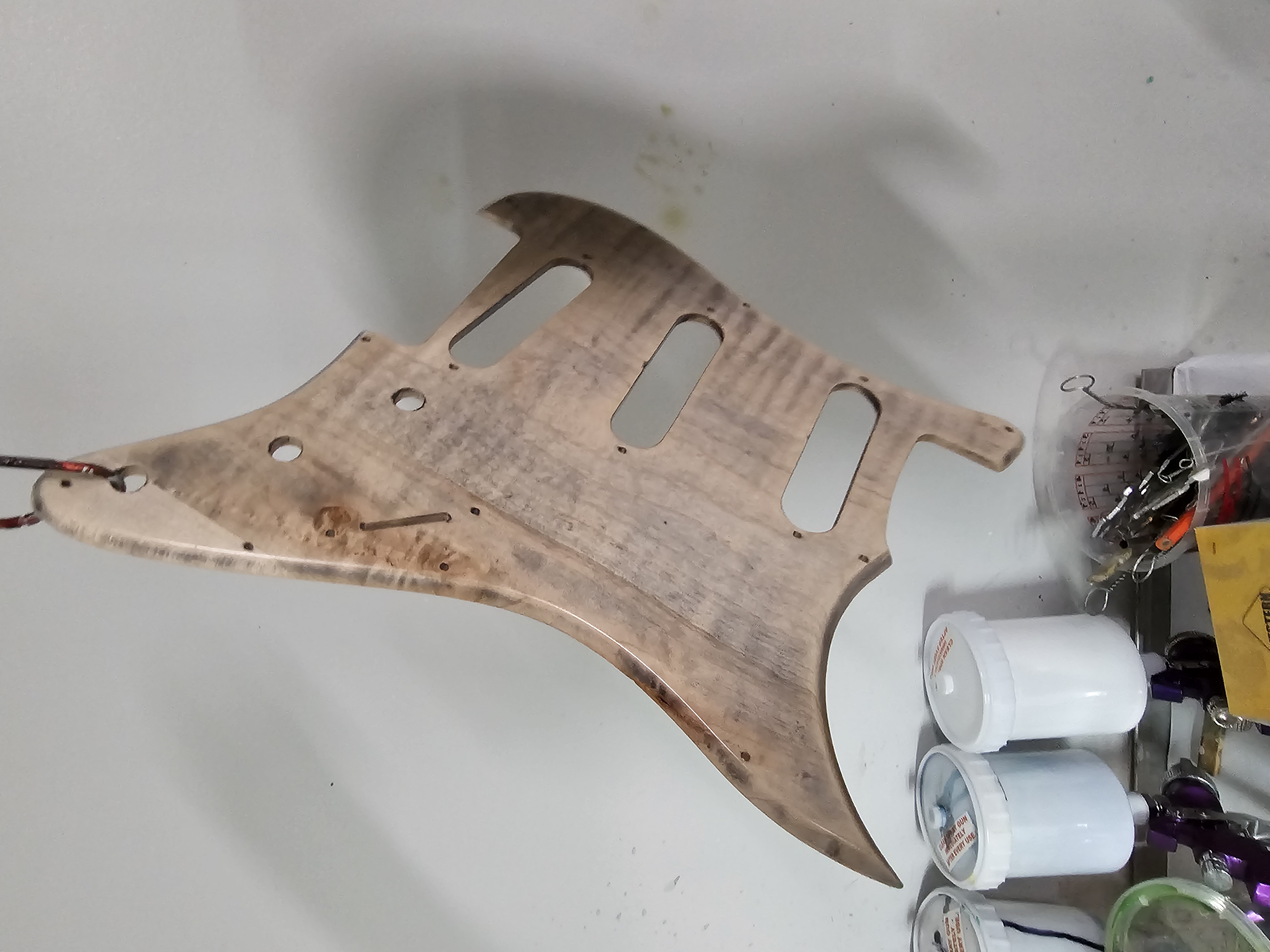A wooden guitar body, unfinished, resting on a white surface, with small containers and tools nearby.