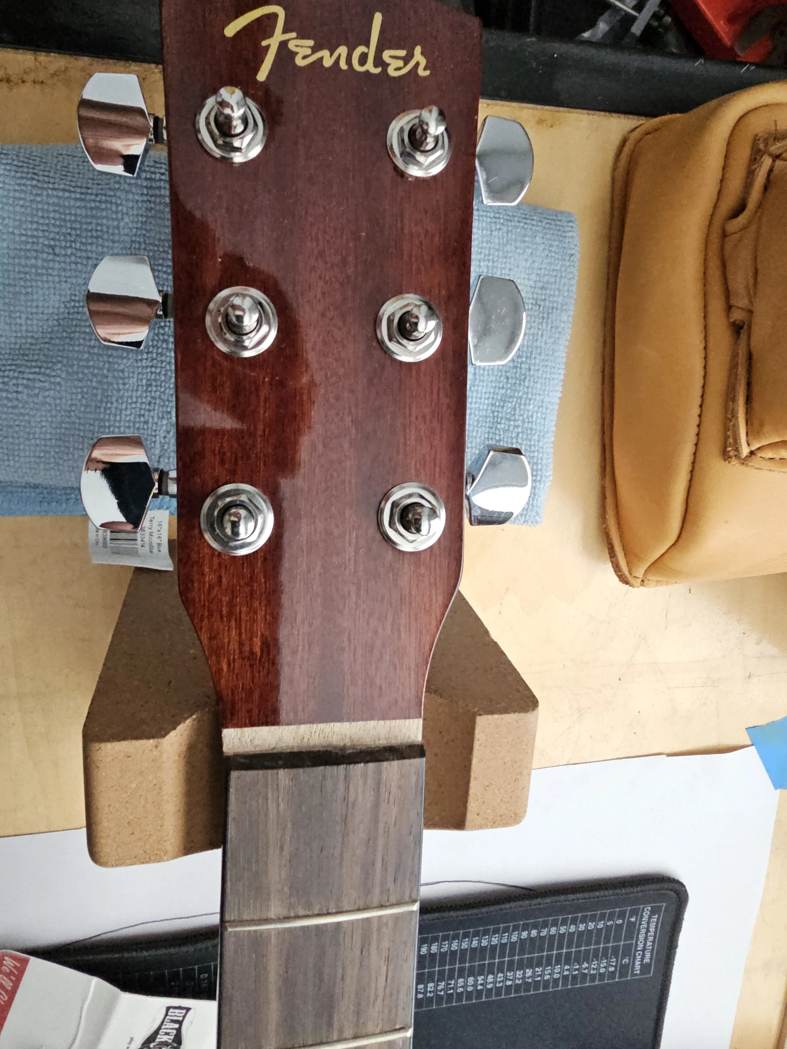 Close-up of the headstock of a Fender guitar with six tuning pegs, on a workbench with a blue cloth and some furniture or cushions in the background.