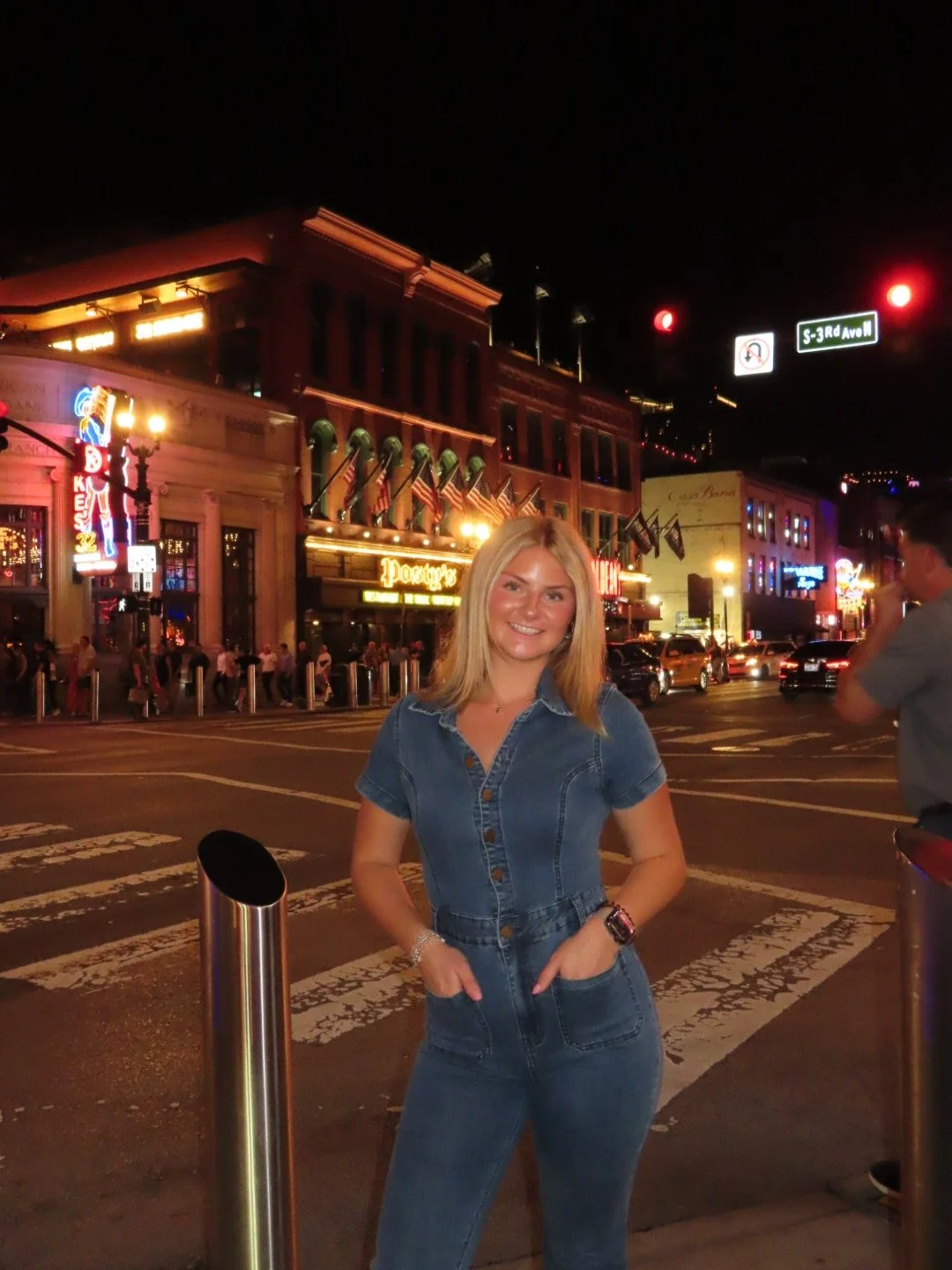 A woman in a denim outfit stands on a city street at night with colorful neon signs and historic buildings in the background.