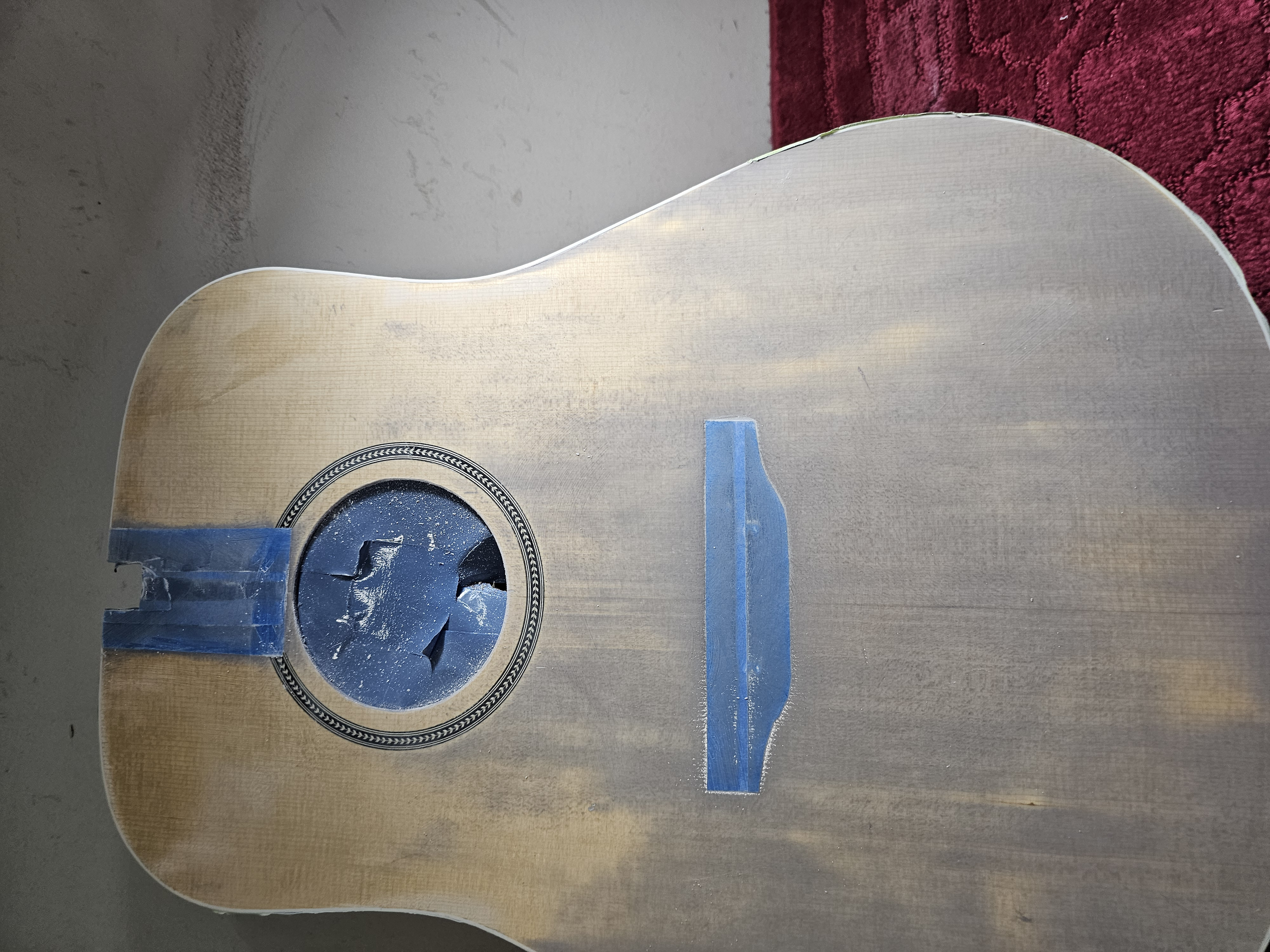 The top of an acoustic guitar body with the sound hole and bridge, taped with blue painter's tape, and a red textured fabric surface partially visible in the background.
