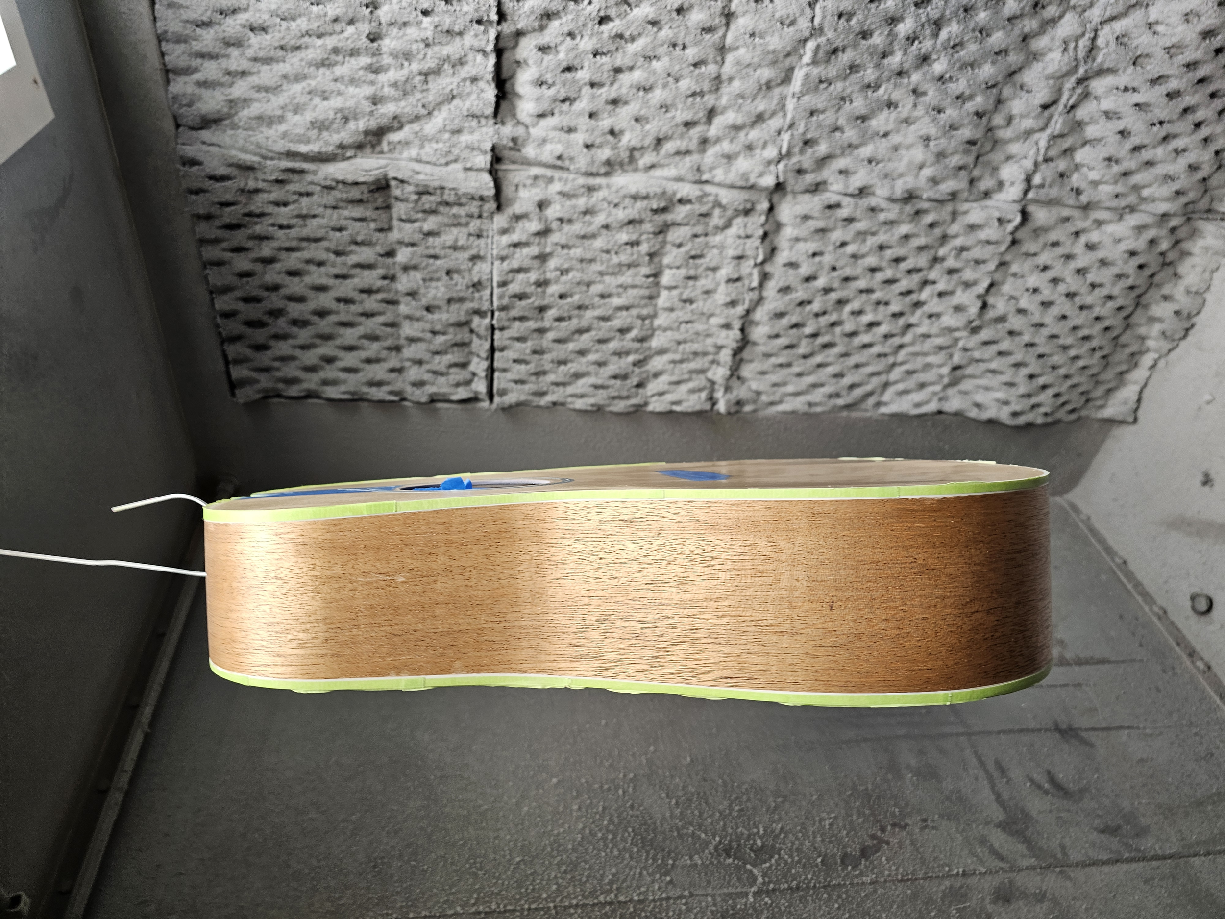 Side view of an acoustic guitar with a wooden body, resting against a gray wall with textured insulation material.