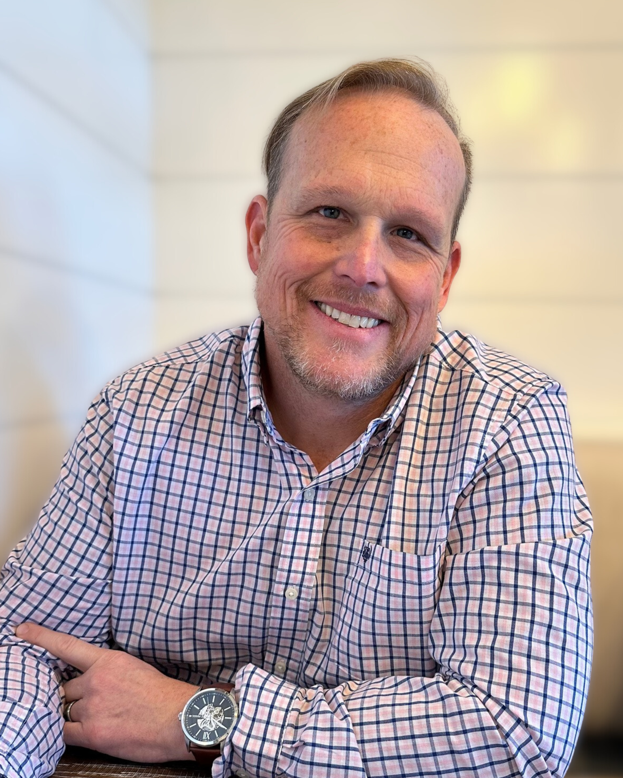 Man in checked shirt smiling at camera, sitting at table with cream-colored wall background.