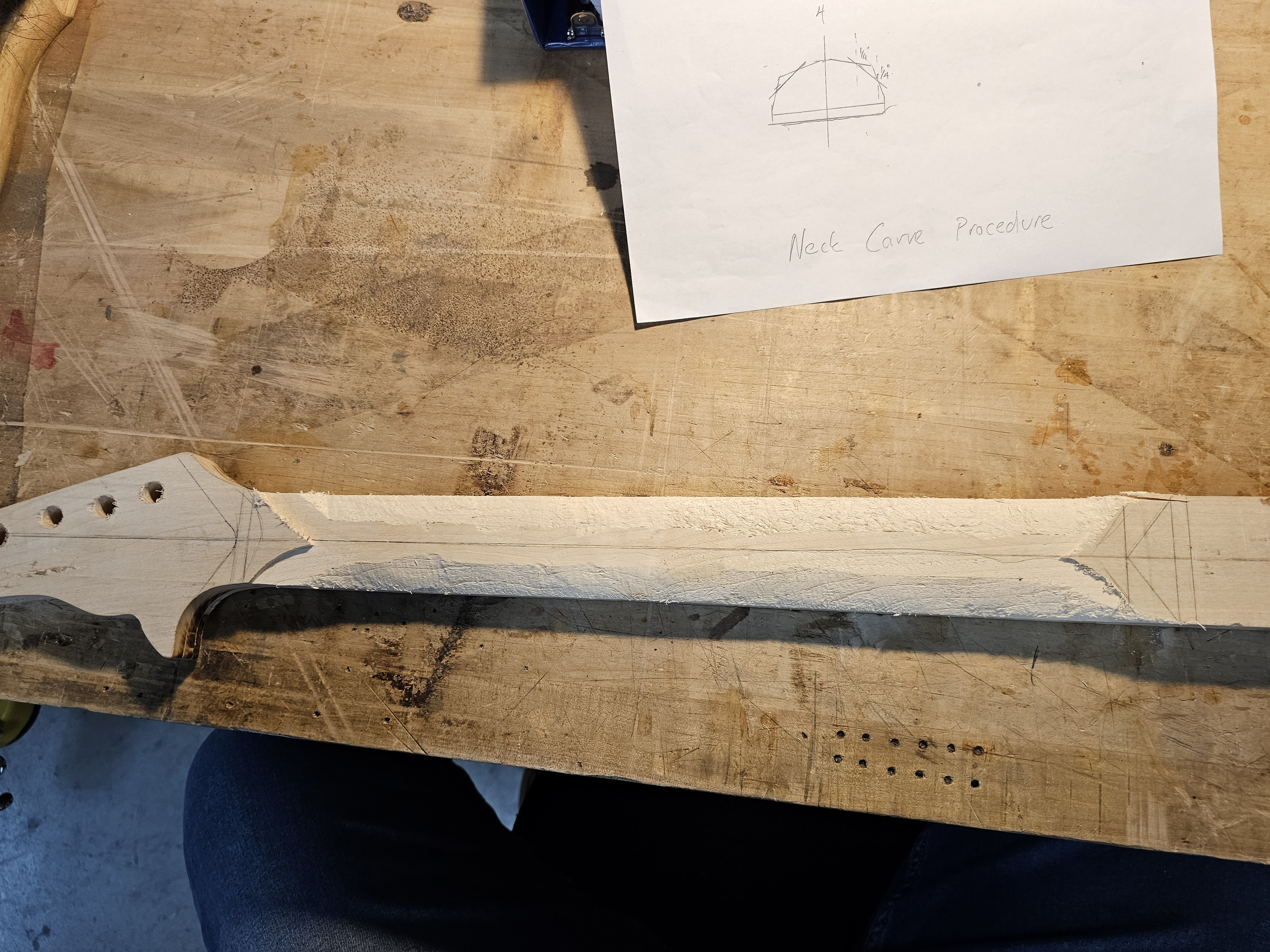 Wooden piece with carved section, dotted hole pattern, and measurement markings, on a workbench next to a paper with a sketch of a semicircular object and notes titled 'Neck Carve Procedure'.