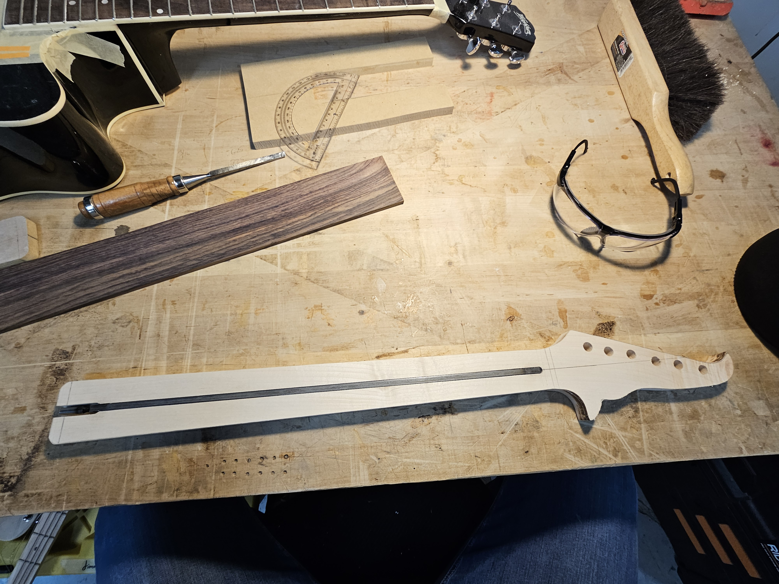 A woodworking workshop table with various tools and materials, including a partially assembled musical instrument neck, a wooden piece, safety glasses, and a protractor.