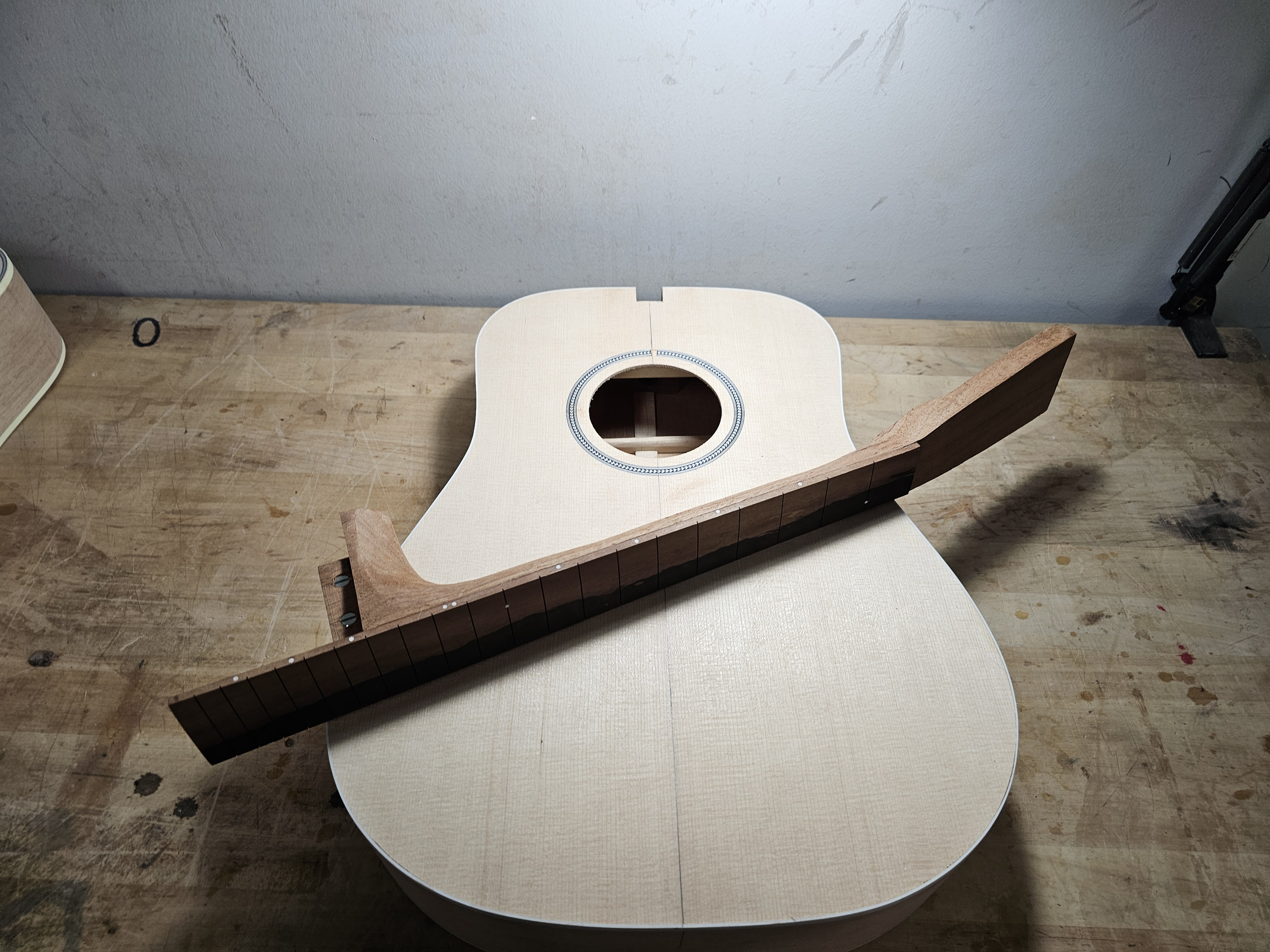 An unfinished acoustic guitar body on a workbench, with a wooden guitar neck placed across it.