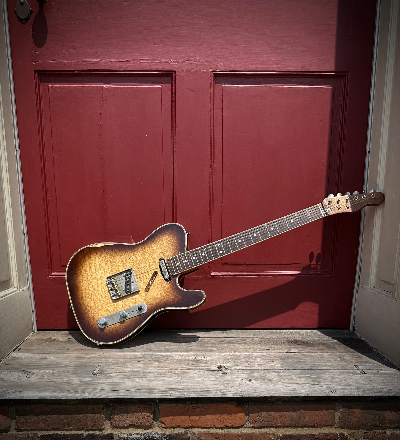 A vintage electric guitar resting on a wooden porch in front of a red door.