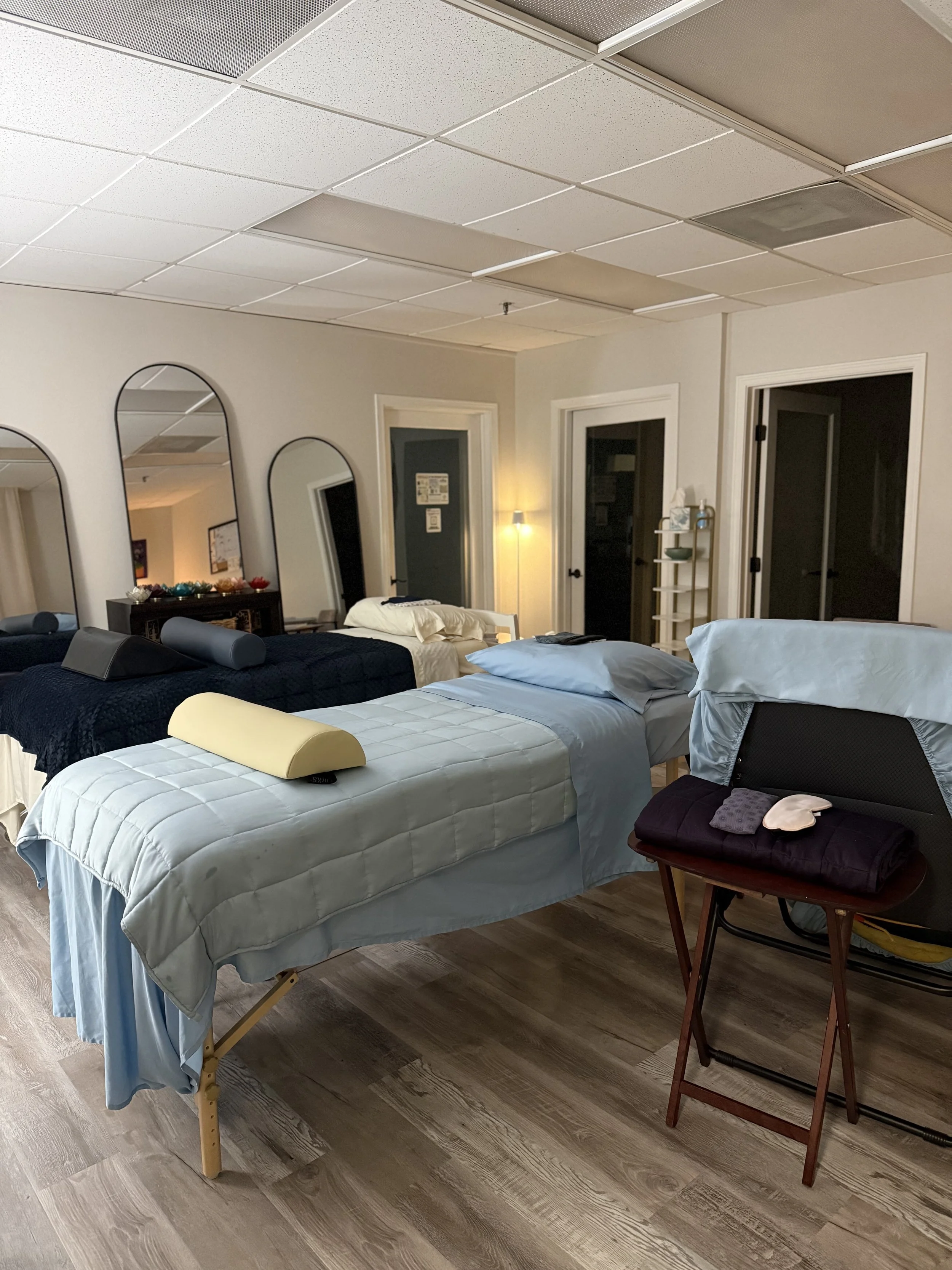 A room with multiple beds, mirrors on the wall, a side table, a wall lamp, and a folding chair with folded towels on it.