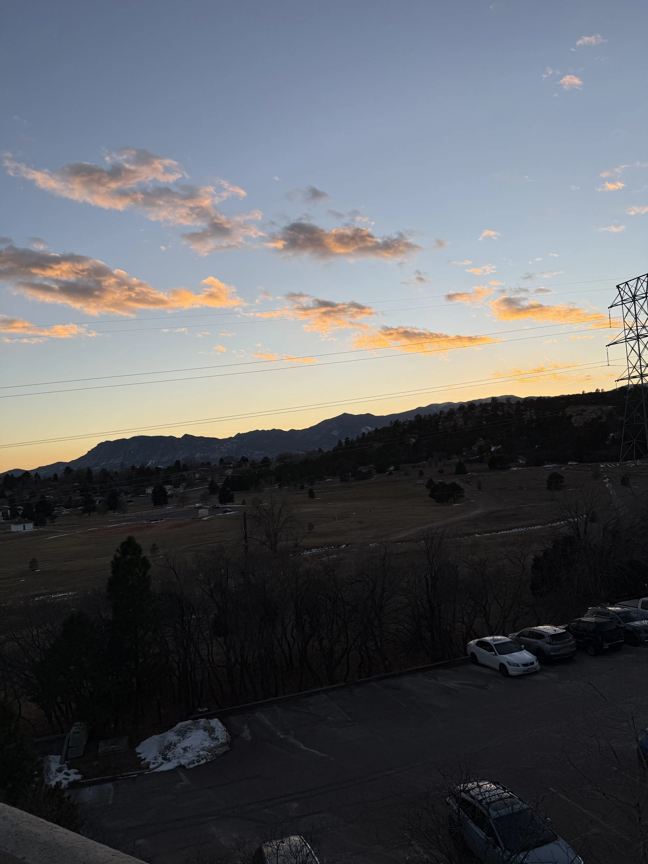 View of mountains and scattered clouds during sunset with power lines and parked cars in the foreground.