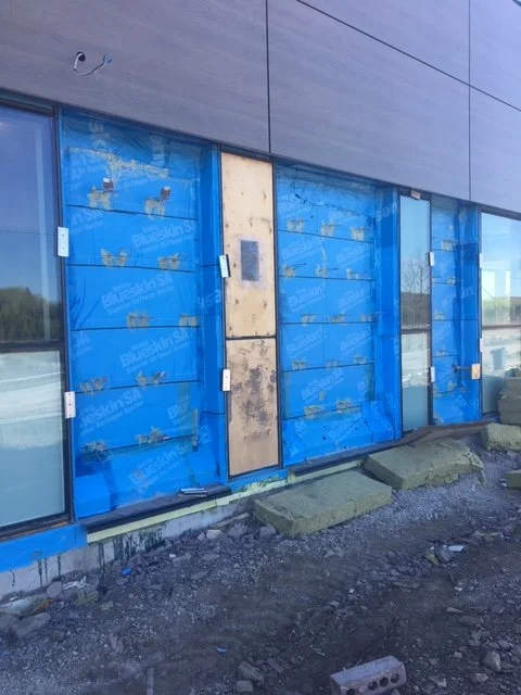 Building under construction with blue protective wrapping on exterior walls, a door covered in dirt, and construction debris on the ground.