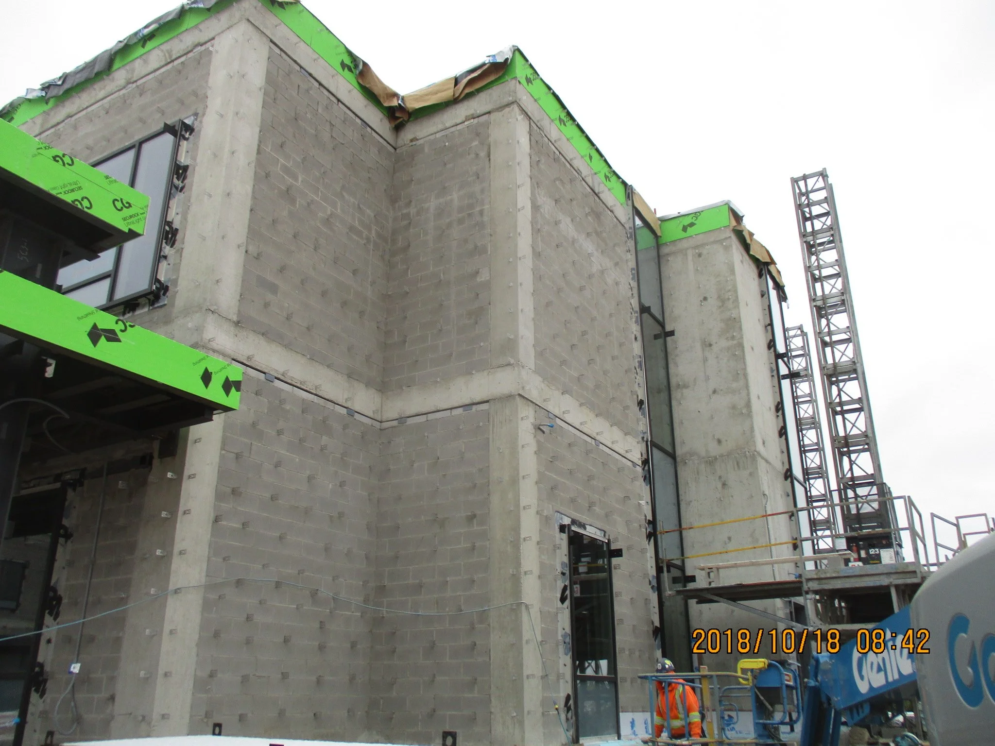 Construction site of a modern building, with concrete walls, large glass windows, and scaffolding on the right side. Construction worker in orange safety vest visible near the bottom right corner.