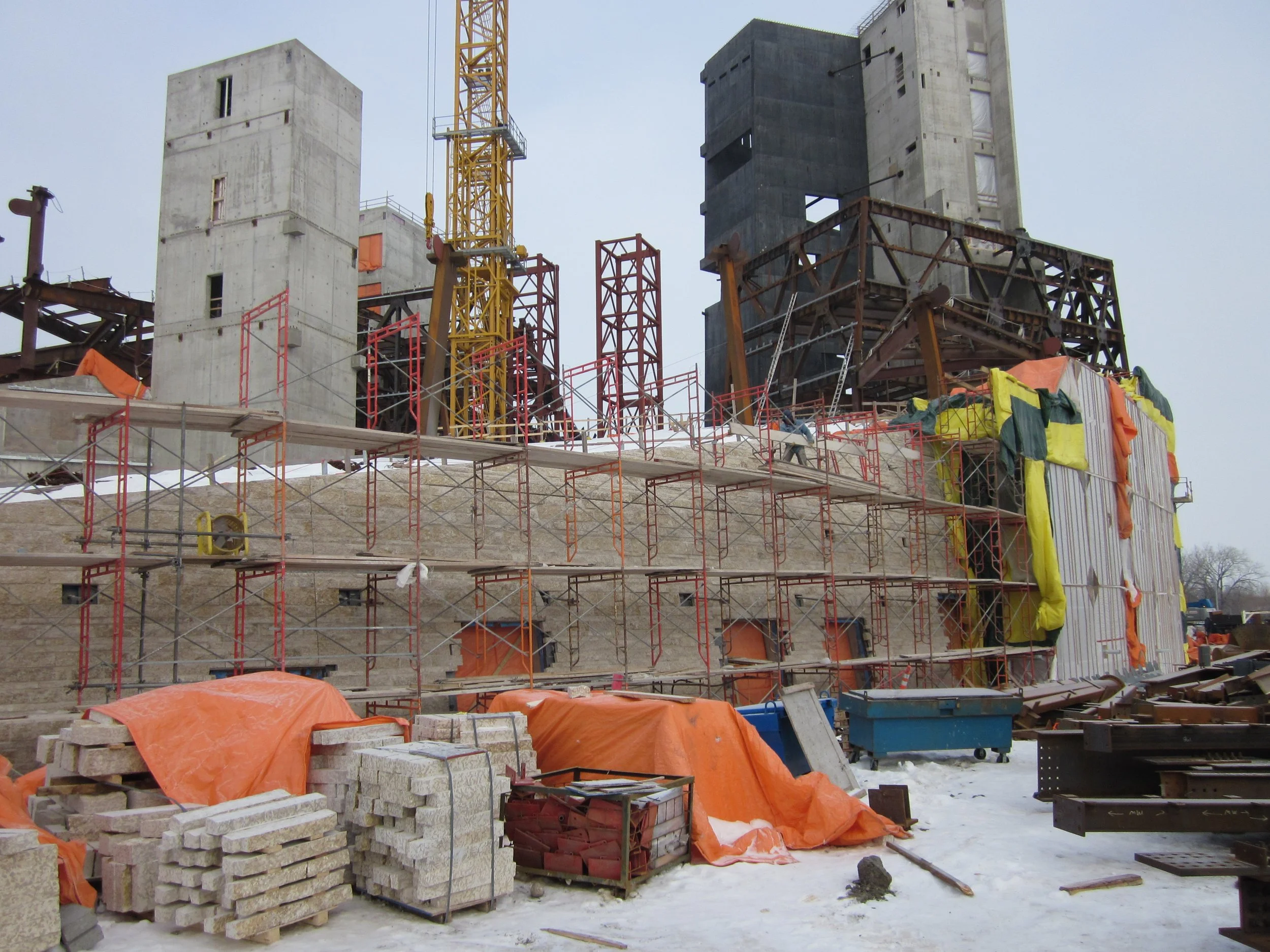 Construction site with scaffolding, concrete structures, building materials, and a yellow crane in a snowy environment.